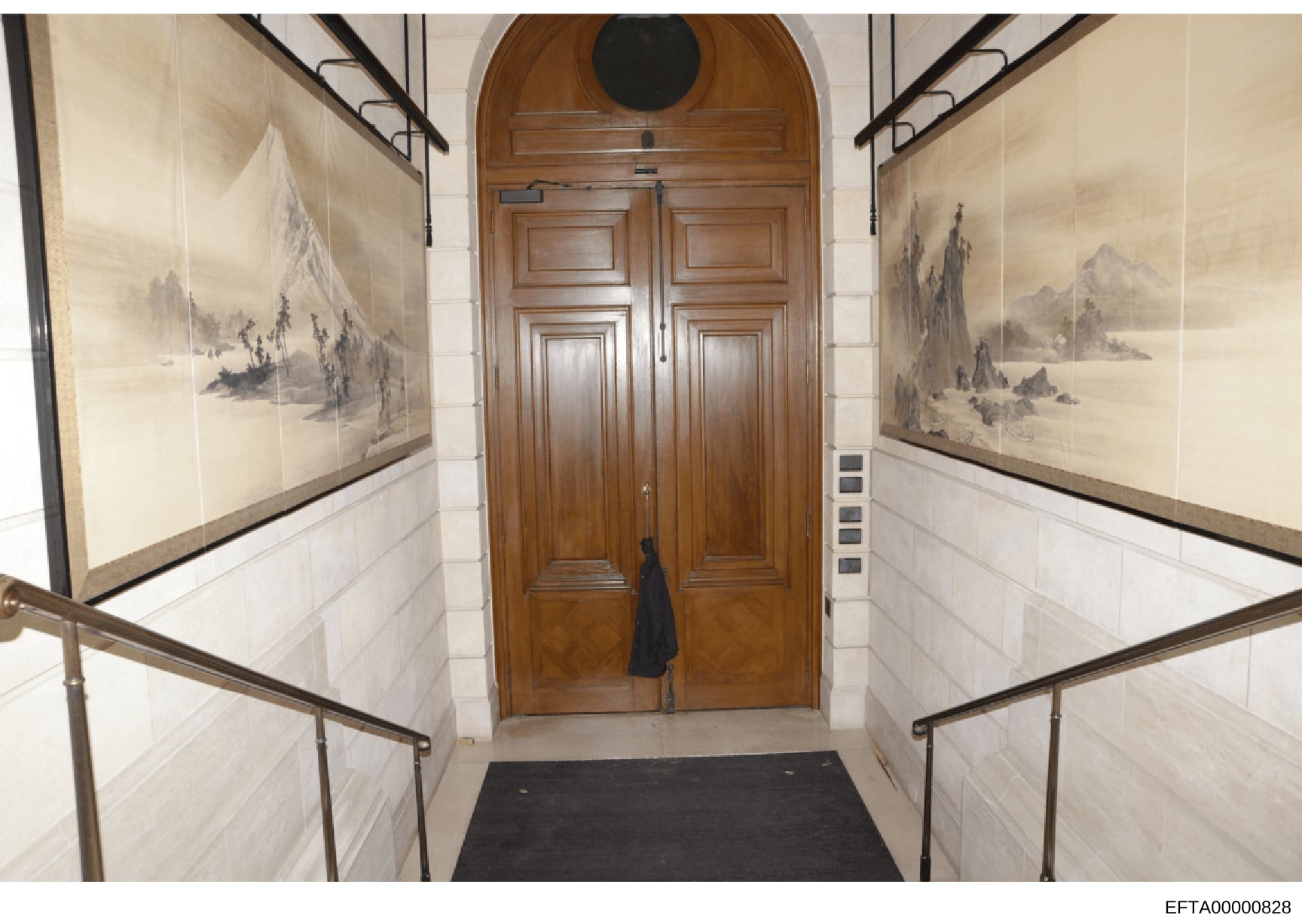 This photograph documents the interior entrance hallway of what appears to be an upscale residential property. The hallway features symmetrically framed landscape artwork on both walls, elegant tile wainscoting, brass handrails, and substantial woode