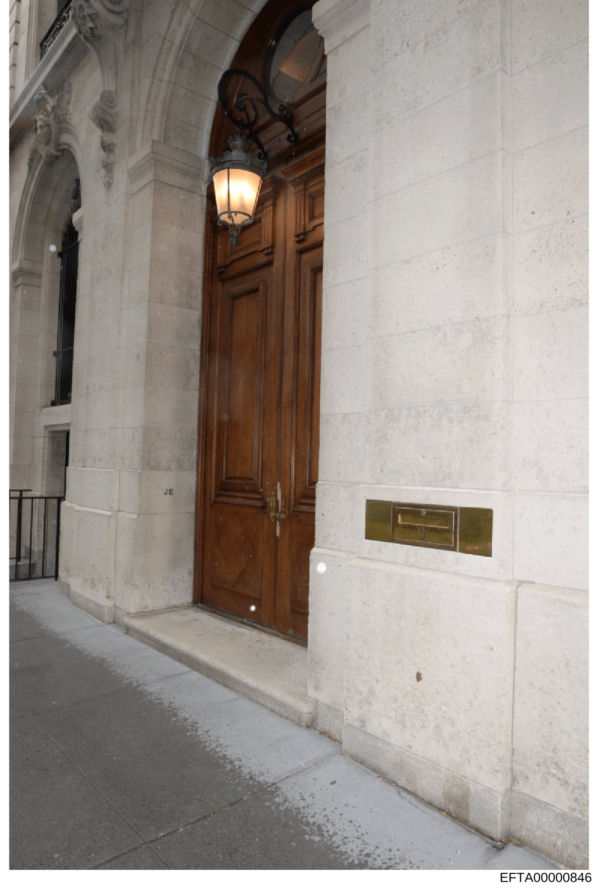 This is a photograph of the exterior entrance to a building, showing a tall wooden double door with brass hardware, white stone facade, decorative wrought-iron lamp fixture, and architectural detailing. The image appears to be documentation of a prop