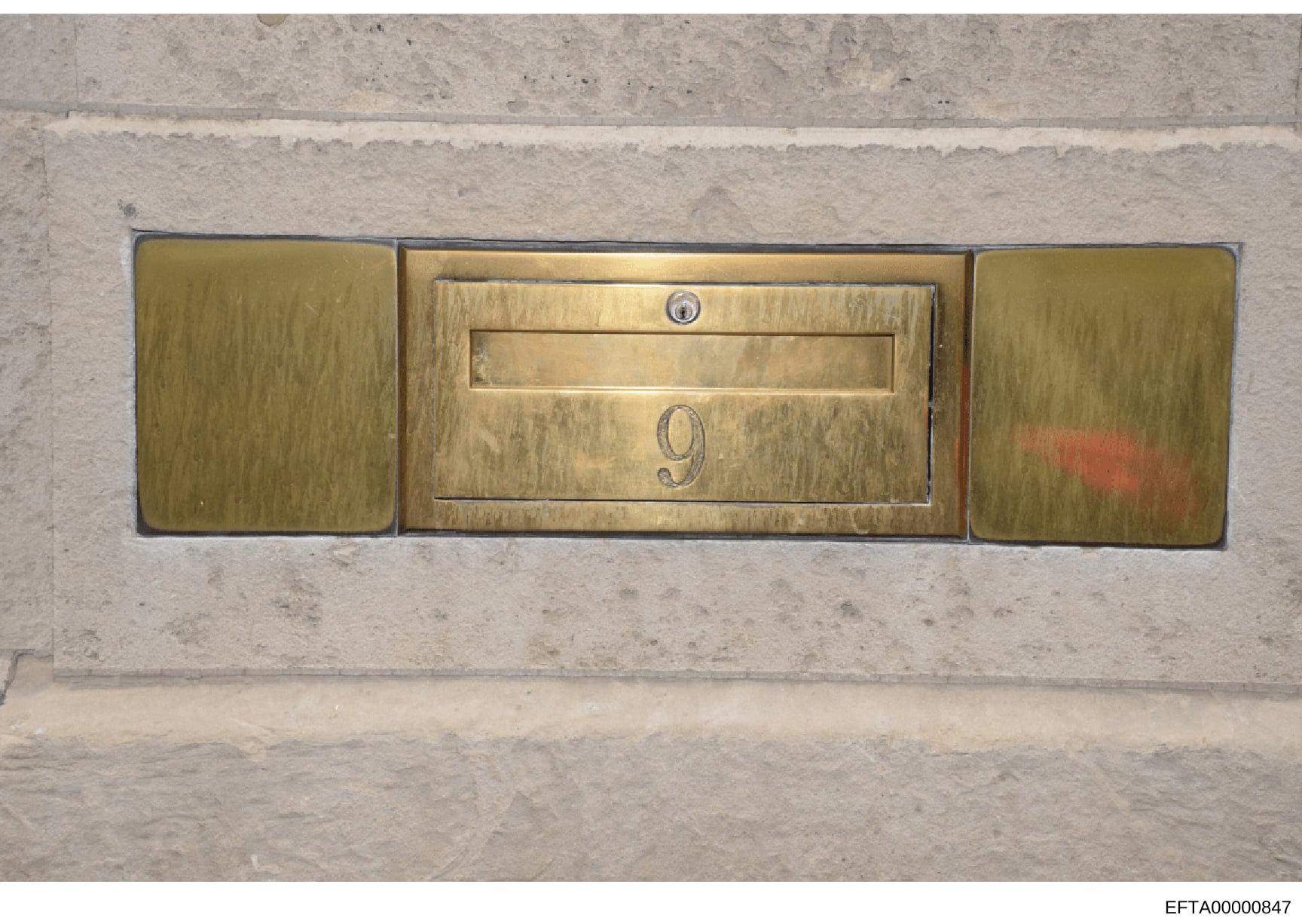 This is a photograph of a decorative brass mailbox mounted on a concrete or stone wall. The three-panel fixture features a central mail slot with the number '9' displayed, flanked by two blank brass panels. The mailbox shows signs of age and patina, 