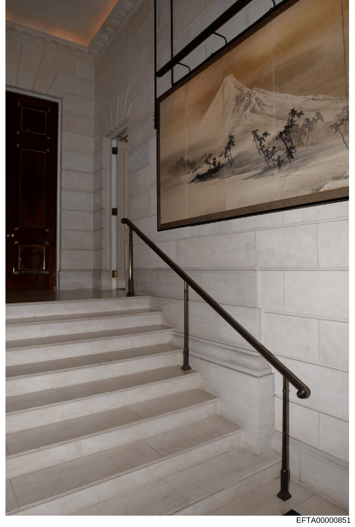 This photograph shows the interior of a stairwell in what appears to be a luxury residential property. The image captures a contemporary design space featuring white marble or stone steps, a dark metal handrail, white subway tile walls, and a large f