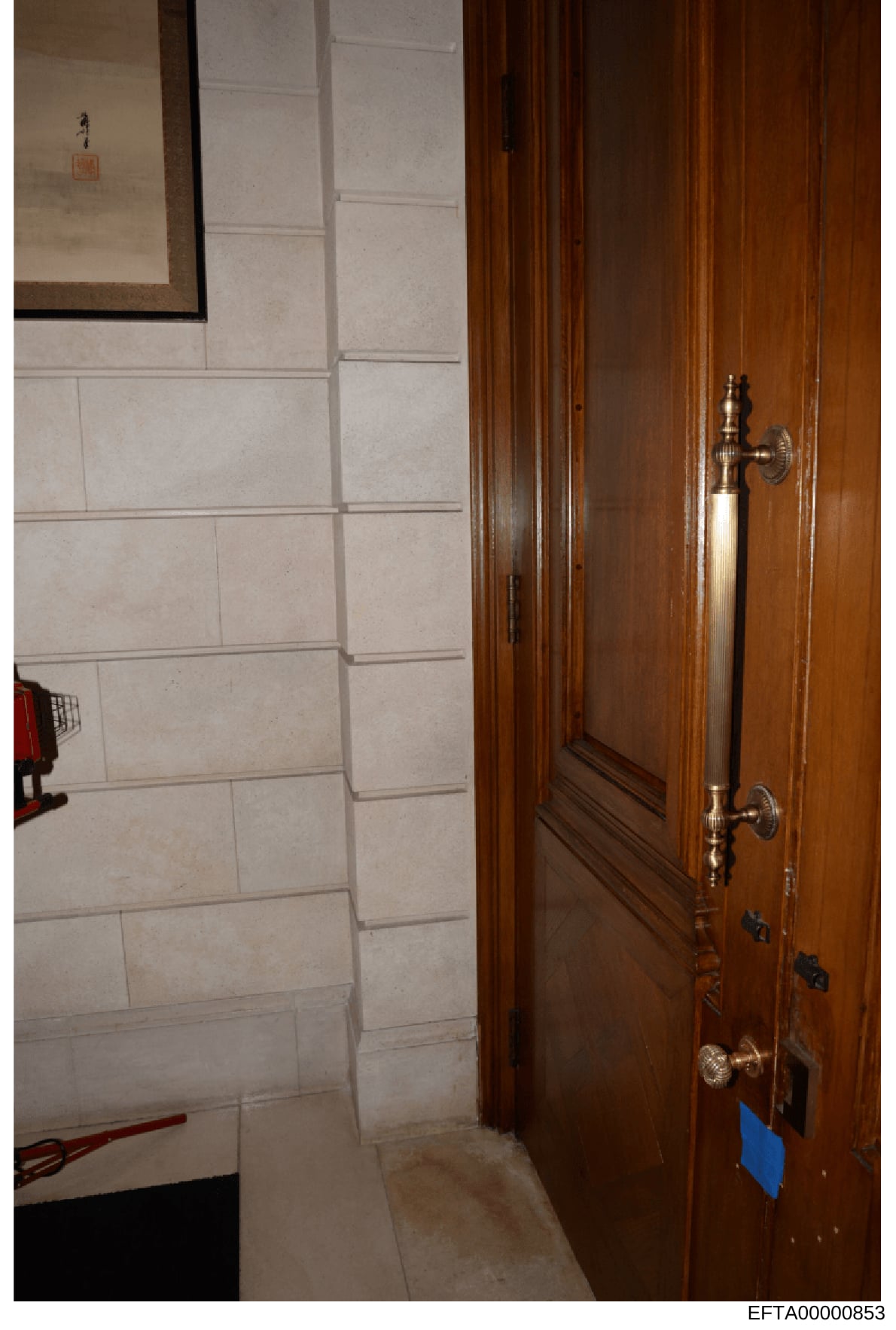 This is an evidence photograph showing the interior of a residential space, featuring a wooden door with brass handles on the right side and cream/beige tile wall paneling on the left. A framed item hangs on the upper left wall. The photo appears to 