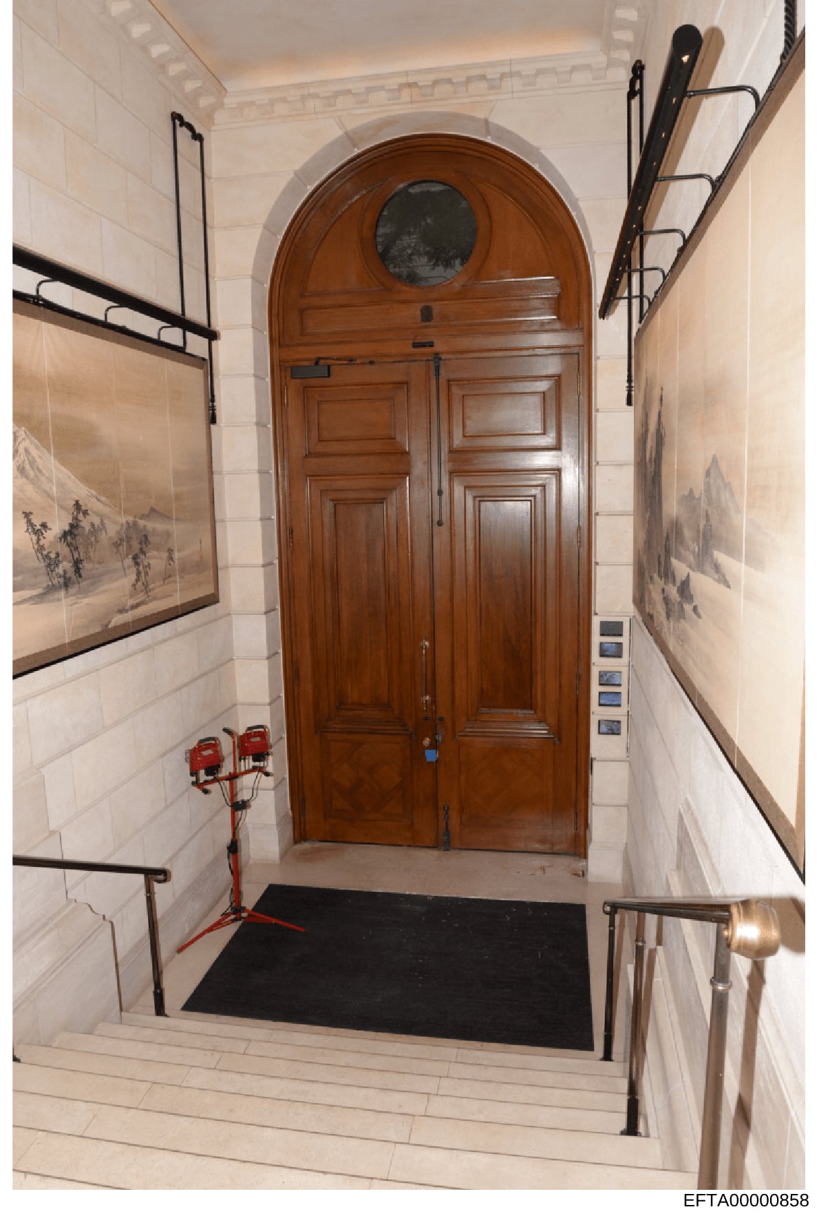 This is a photograph of an upscale residential building's interior entryway or foyer, featuring a distinctive arched wooden double door with a circular transom window. The space shows elegant architectural detailing including decorative framed artwor