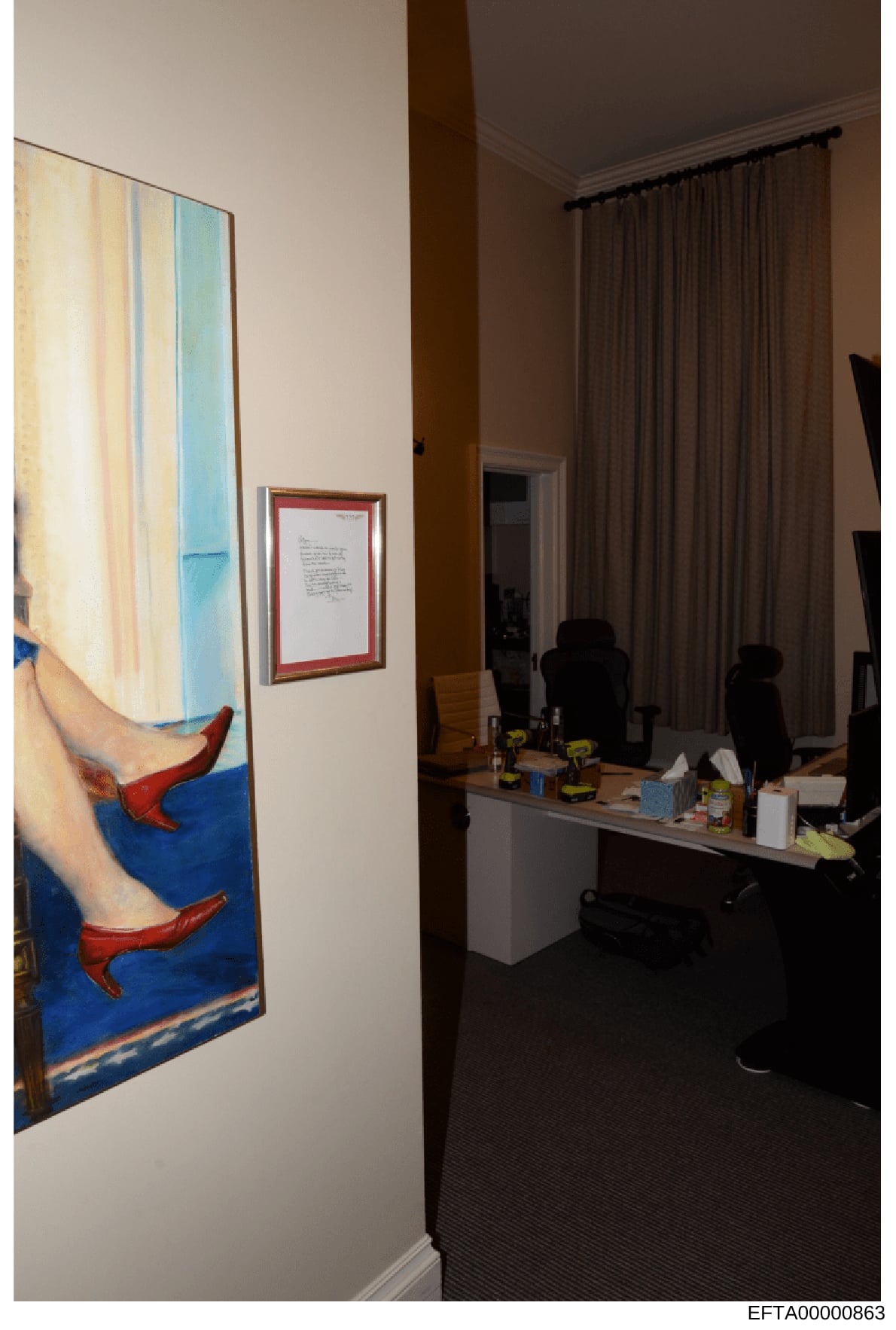 This is a photograph of an interior office or residential space showing a modern workplace setup with contemporary artwork displayed on the walls. The image captures a desk area with office equipment, curtained windows, and framed art including what