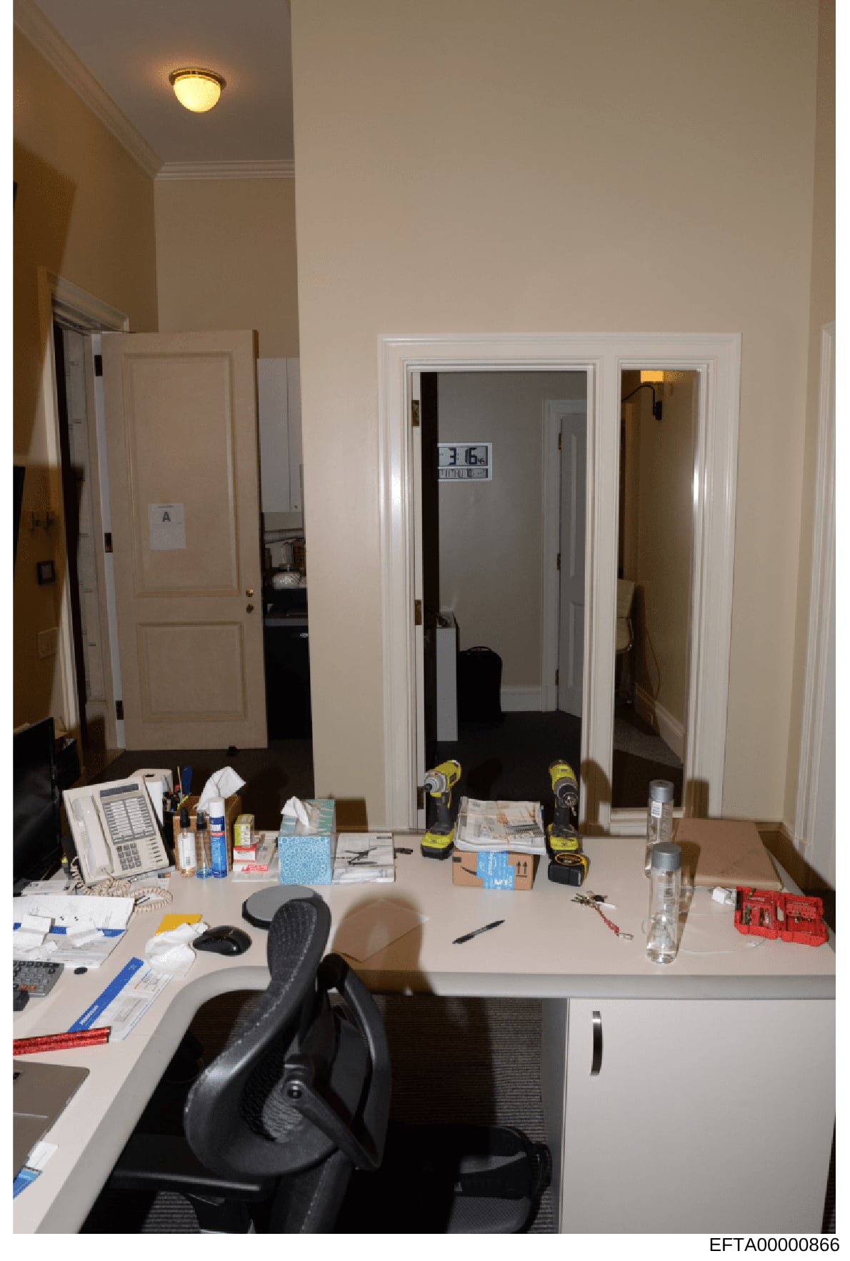 This photograph shows an interior office or workspace with a desk setup containing various tools, documents, and equipment. The room features white walls, cream-colored trim, and professional furnishings including a black office chair. Multiple doors