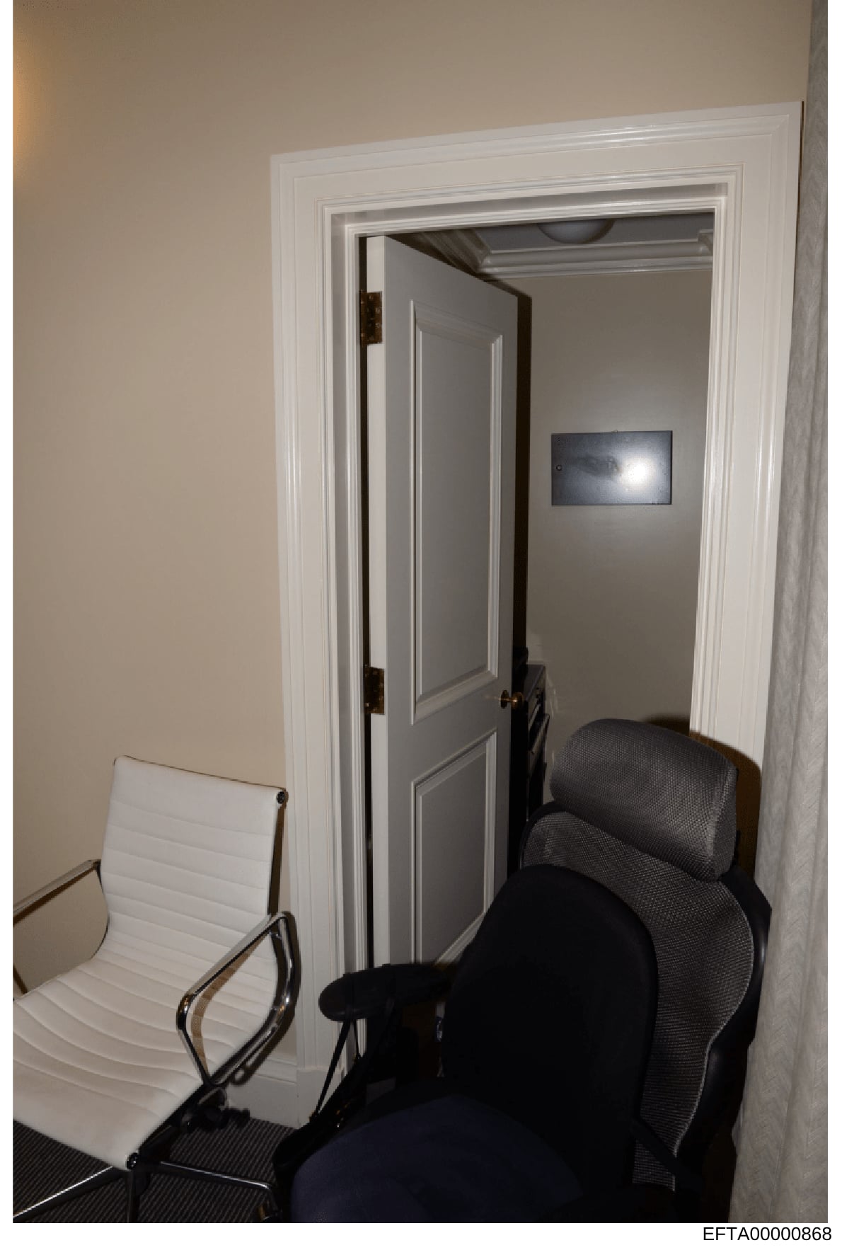 This is a photograph of an interior office or waiting area showing a small room with modern furnishings. The image depicts two chairs (one white, one black mesh office chair) positioned in front of an open doorway leading to an adjacent room with gra