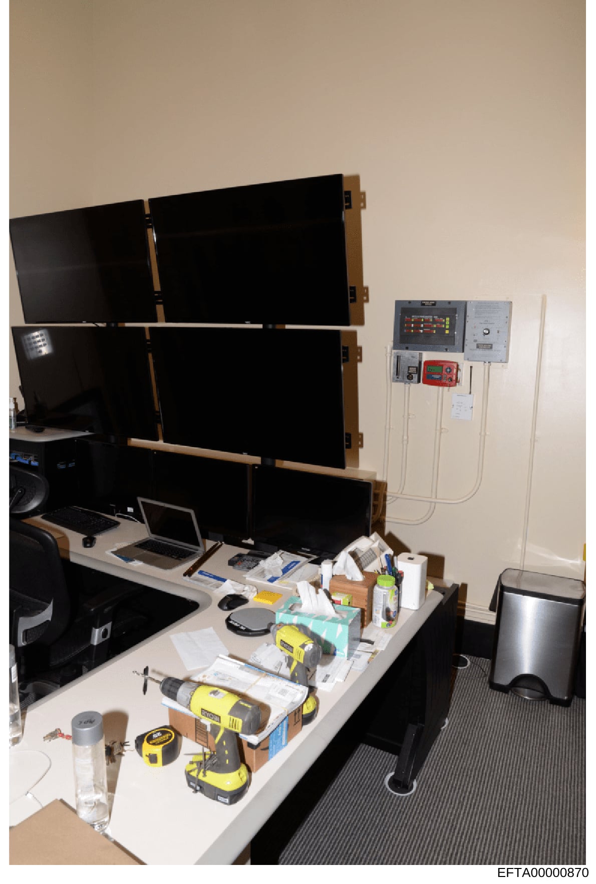 This photograph shows a control room or monitoring station setup with six large black display screens arranged in a grid pattern, mounted on a wooden frame. Below are laptops, various electronic monitoring equipment, and tools scattered on a desk. Th