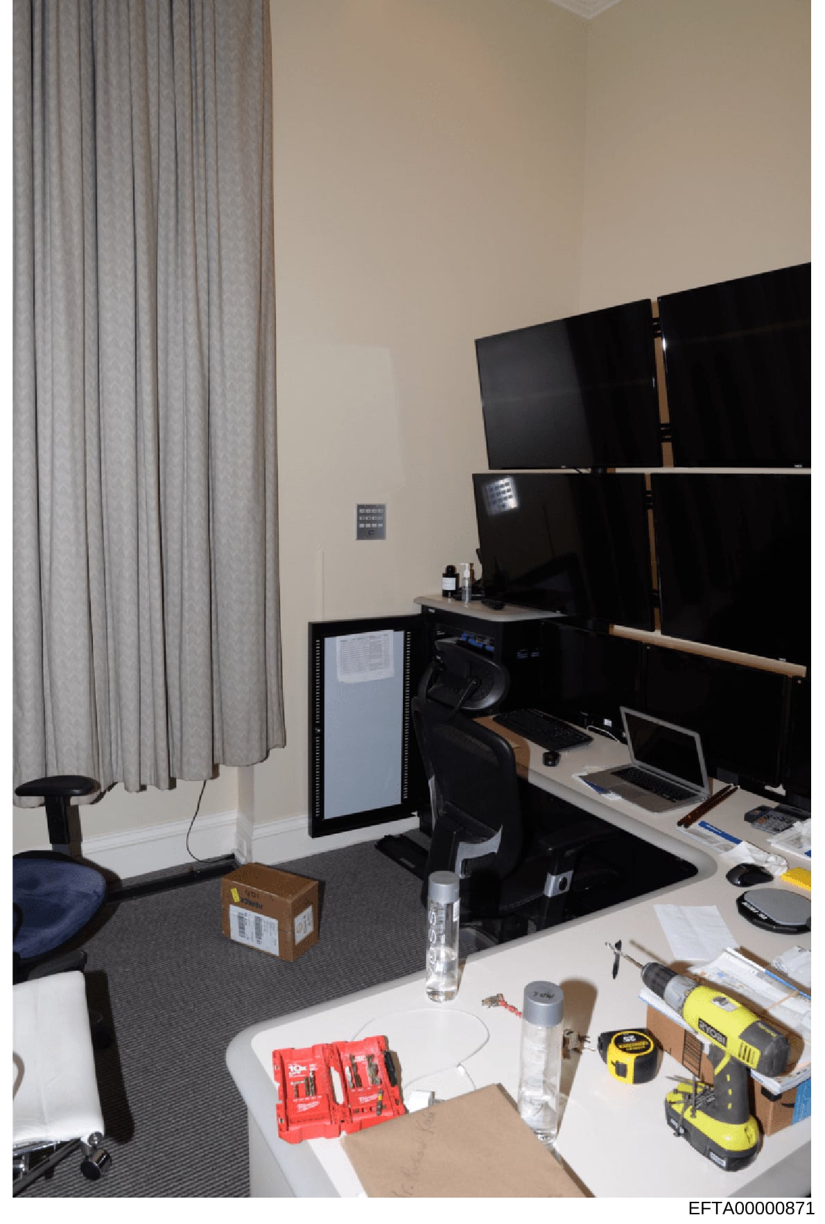 This is a photograph of an interior office or workspace showing a computer workstation with multiple monitors, desk equipment, tools, and furnishings. The image appears to be evidence documentation from the investigation, capturing the physical setup