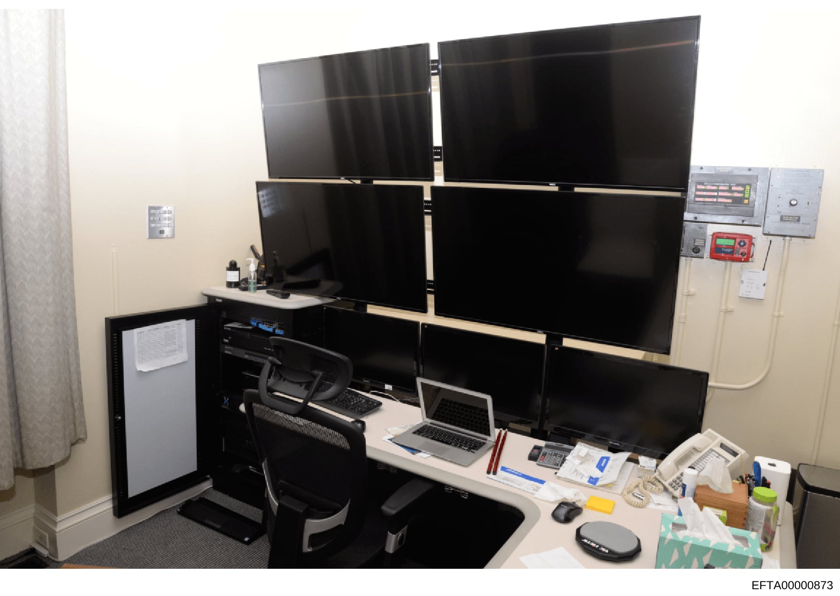 This photograph shows a sophisticated multi-monitor workstation setup with six display screens arranged in a 3x2 configuration, a laptop, and various office equipment. The setup appears designed for monitoring or surveillance purposes, with control p