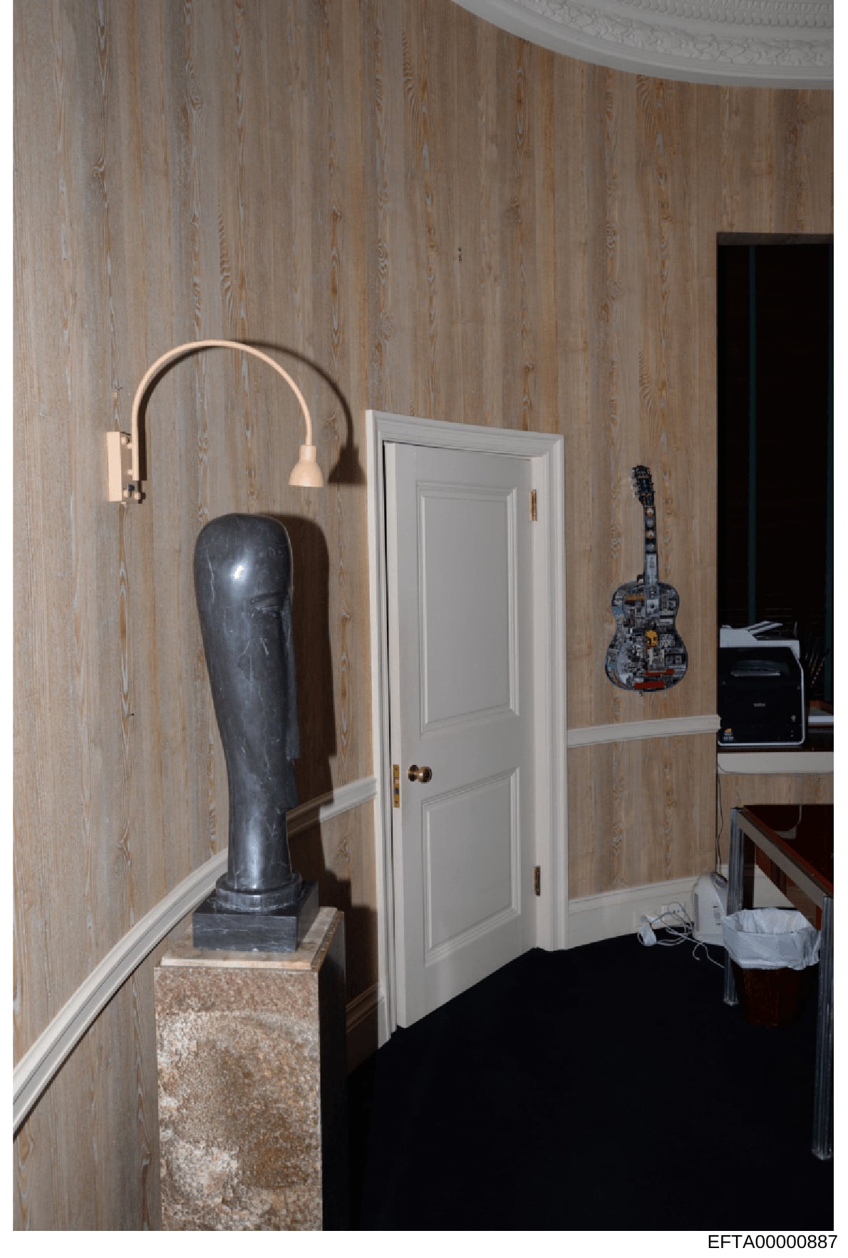 This is a photograph of an interior room showing decorative elements and furnishings, including a dark sculptural bust on a stone pedestal, a wall-mounted arc lamp, a decorative guitar wall hanging, and a white wooden door. The image appears to be ev