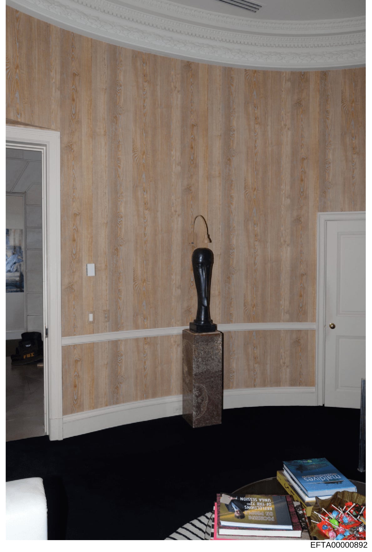 This is a photograph of an interior room featuring curved wood-grain wallpaper walls with white trim, a black sculpture on a stone pedestal, and furnishings including books and decorative items. The image appears to be from a property owned or contro