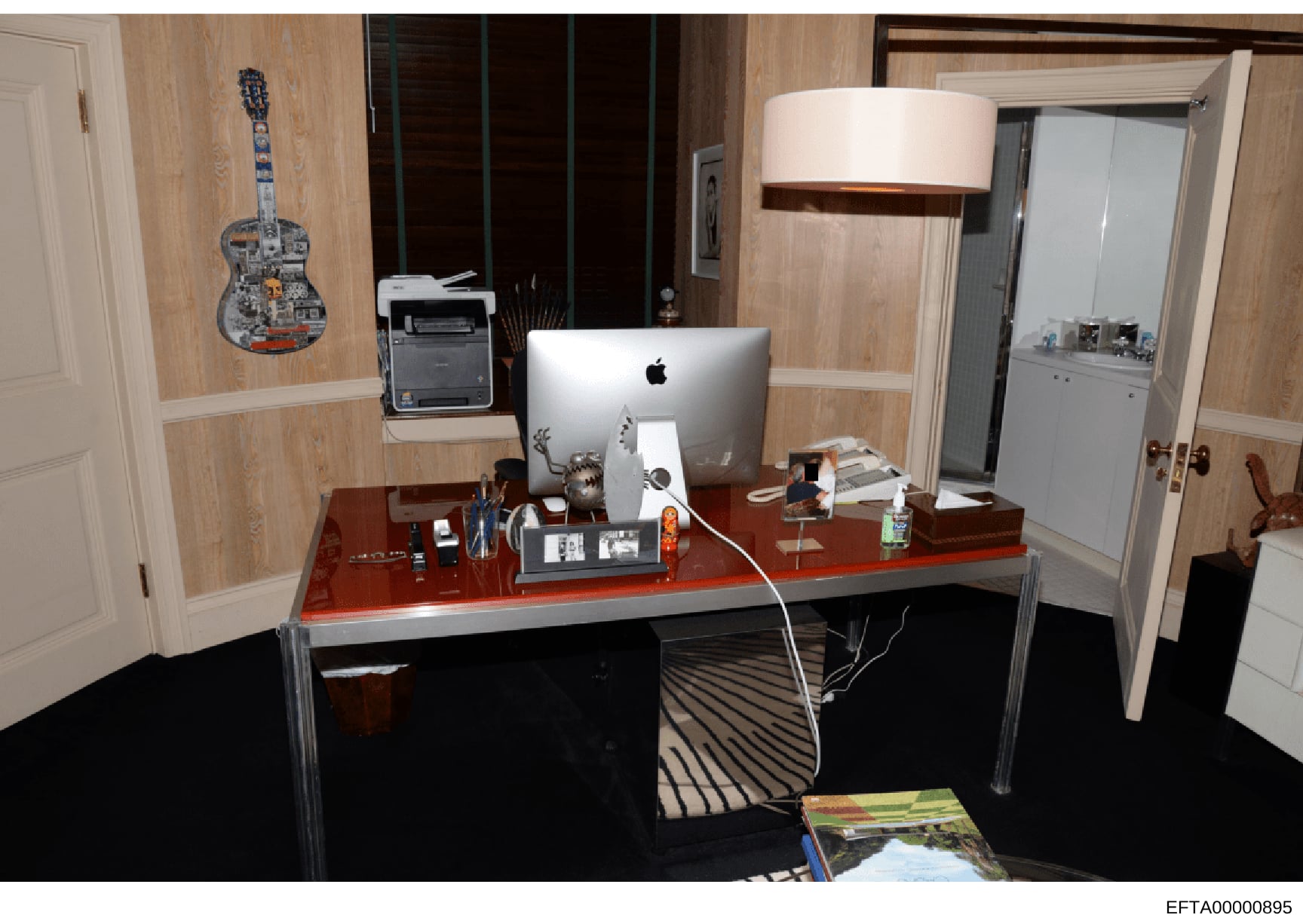 This photograph shows an interior office or study space featuring a wooden desk with computer equipment, decorative artwork, and personal items. The room contains wood-paneled walls, white doors, a framed guitar artwork, and various furnishings. The