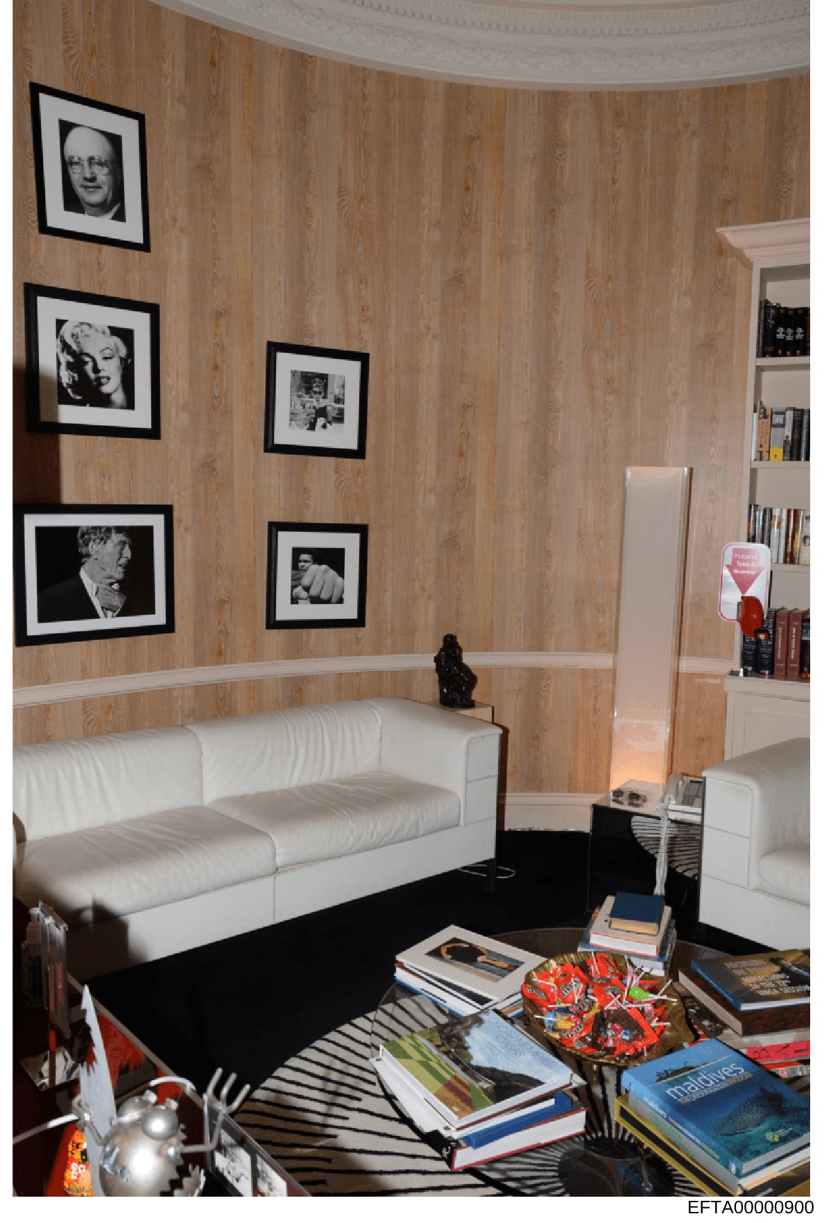 This photograph documents an interior room, likely from one of Jeffrey Epstein's properties, featuring wood-paneled walls with framed black-and-white portrait photographs, white upholstered seating, built-in shelving, and various decorative items and