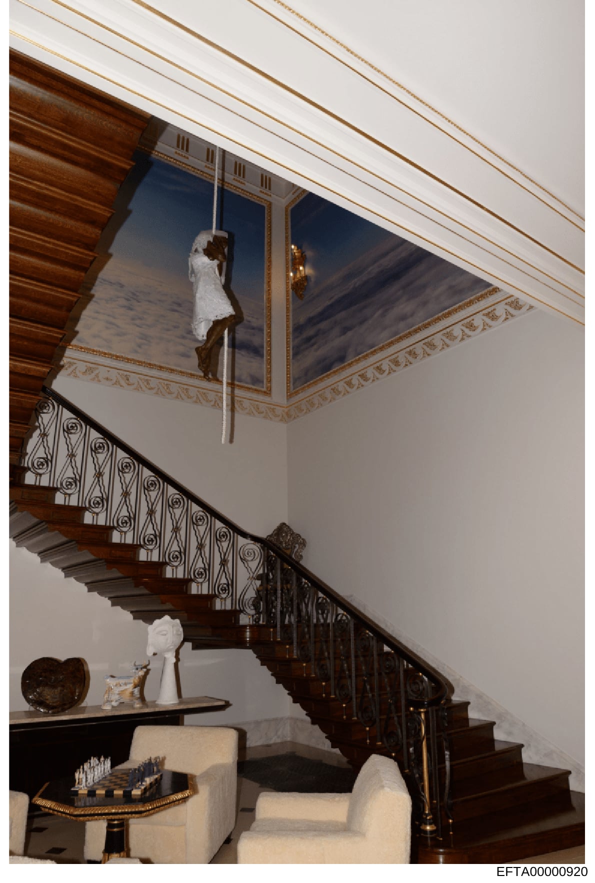 This photograph shows the interior of an upscale residence featuring a curved wooden staircase with ornate wrought-iron railings, cream-colored walls, and a striking trompe-l'oeil ceiling mural depicting a figure on a rope against a sky background. T