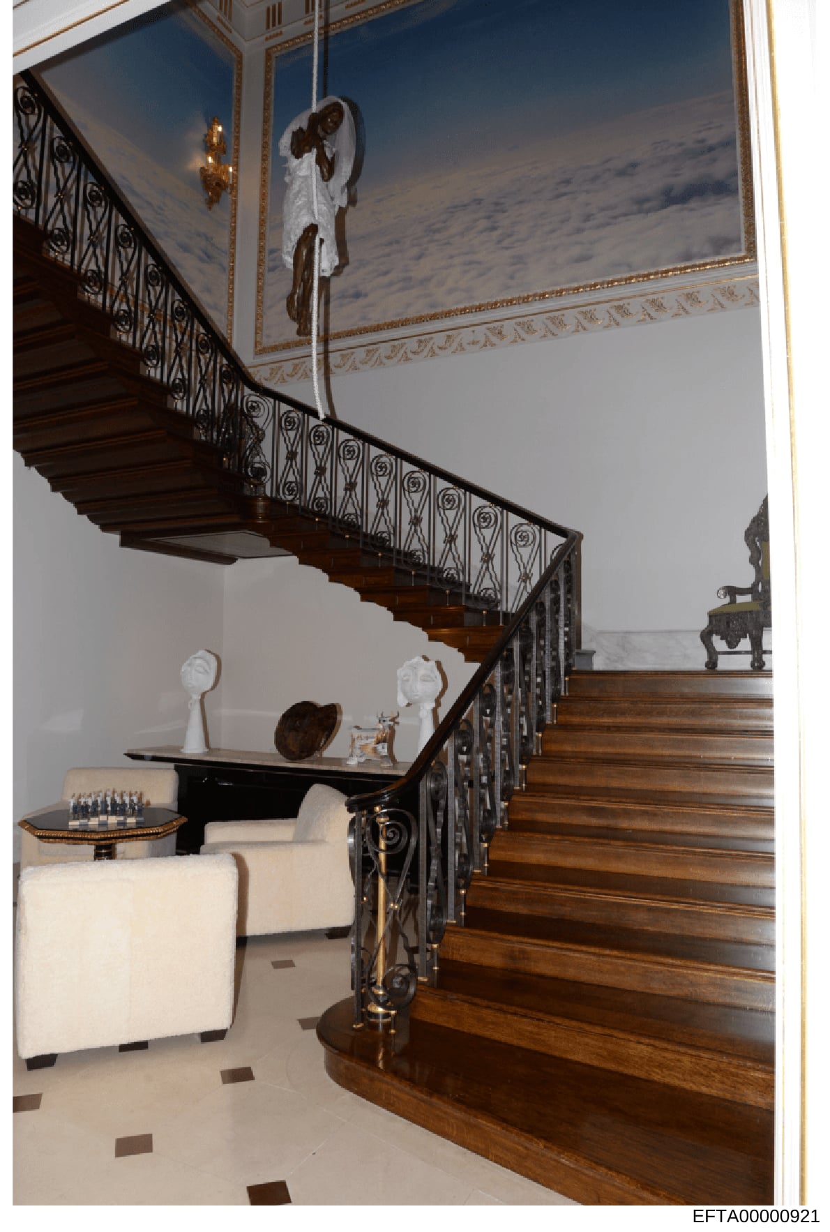 This is a photograph of an elegant interior staircase featuring ornate wooden railings, a sweeping curved design, and decorative artwork on the ceiling. The space includes classical sculptures, fine furnishings, and architectural details consistent w
