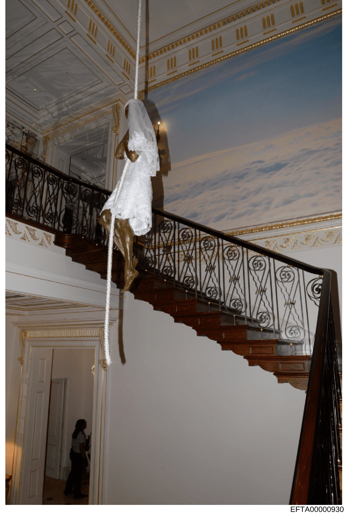 This photograph shows the interior of a luxurious residence featuring an elaborate spiral staircase with decorative wrought iron railings. A white cloth or fabric is draped from the ceiling to a brass fixture, suspended above the staircase. The space