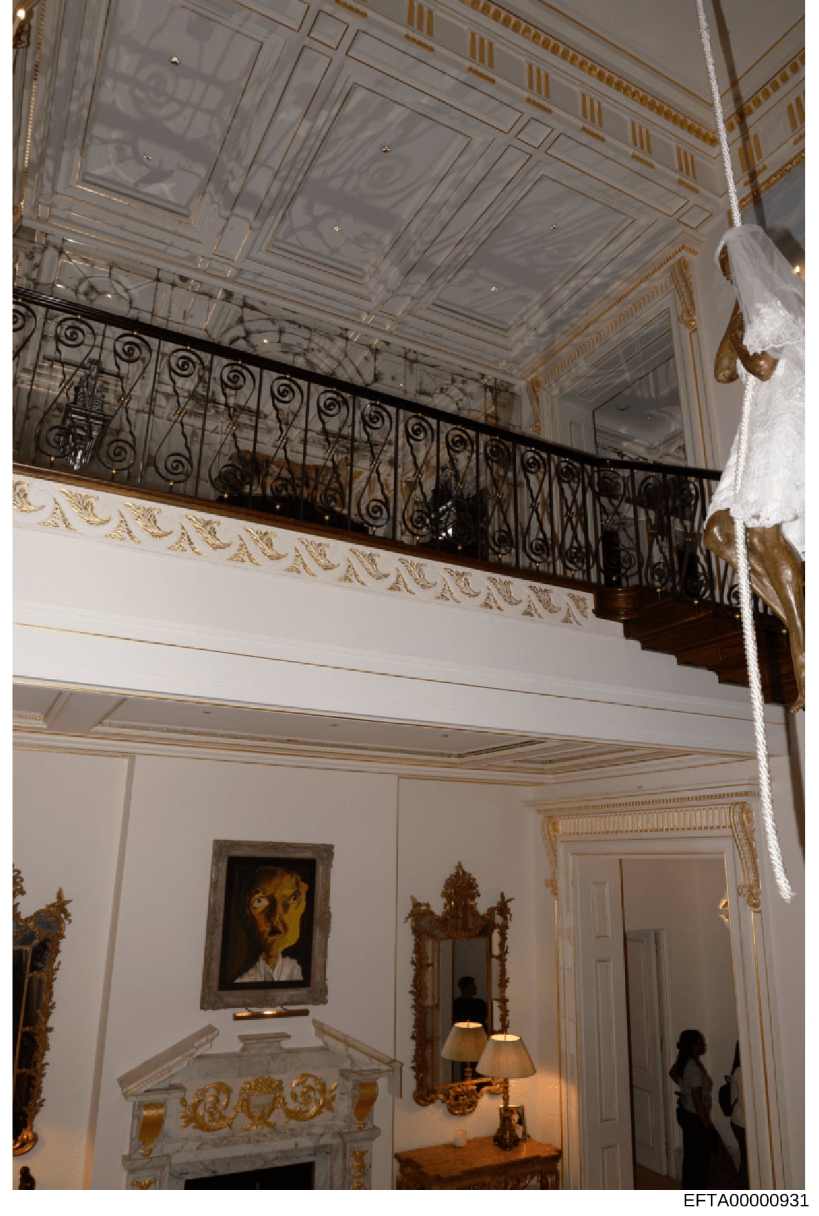 This photograph documents the interior of an opulent residential space, featuring a two-story room with neoclassical architectural elements including decorative ceiling panels, ornamental wrought iron balcony railings, gilt trim, and marble fireplace