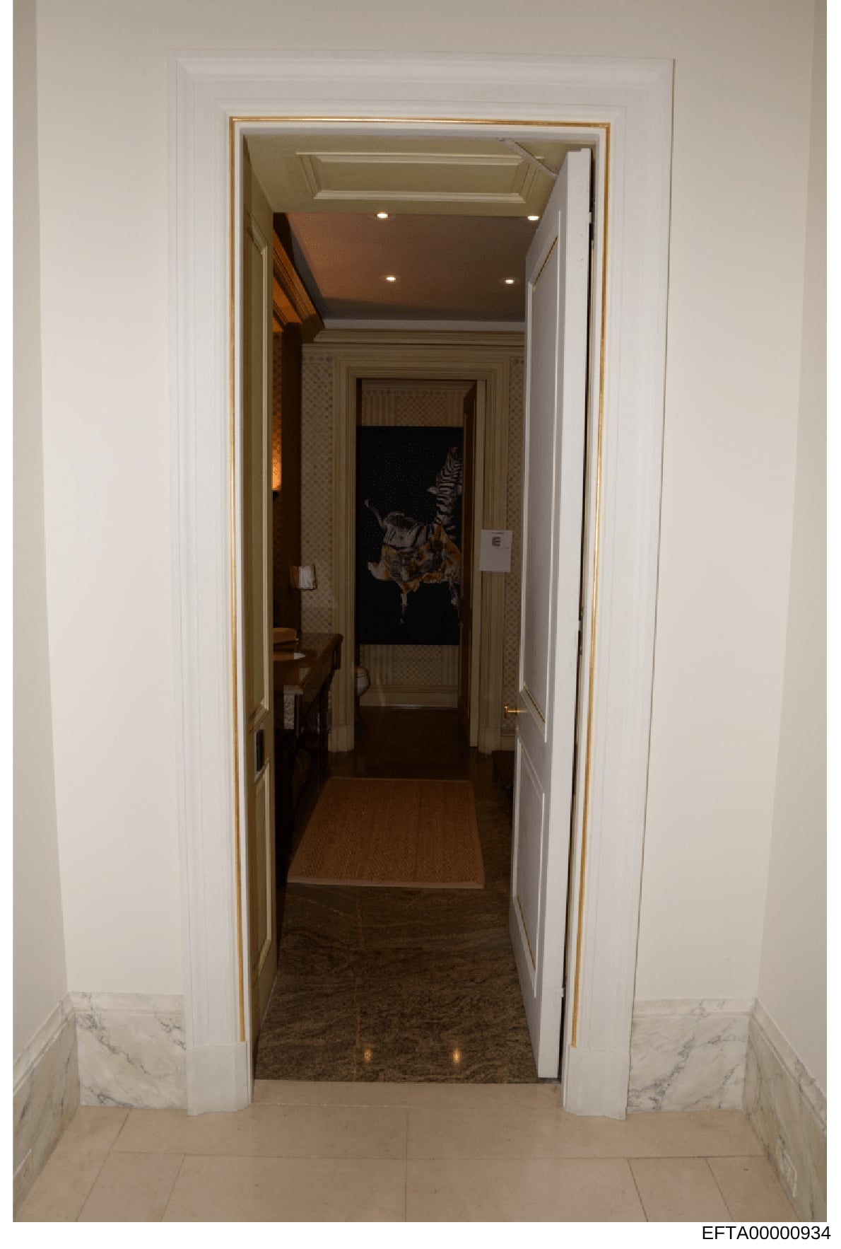 This is an evidence photograph showing an interior hallway or corridor in what appears to be a luxury residential property. The image depicts an elegant entrance with white walls, a gold-trimmed doorway frame, marble baseboards, and recessed lighting
