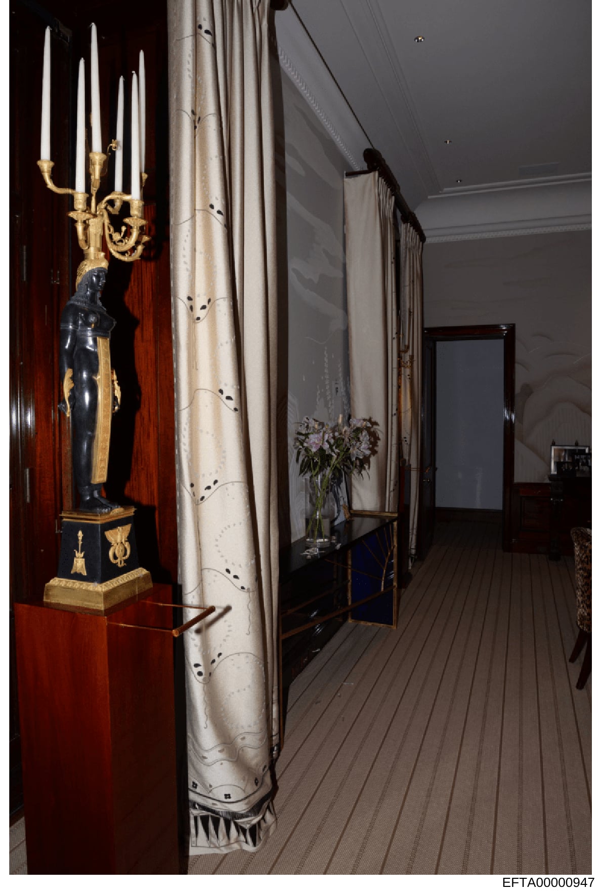 This photograph shows an interior hallway or corridor in what appears to be a luxury residence or yacht. The space features a tall decorative candelabra with gold gilt and white candles mounted on a red lacquered pedestal, accompanied by a black scul