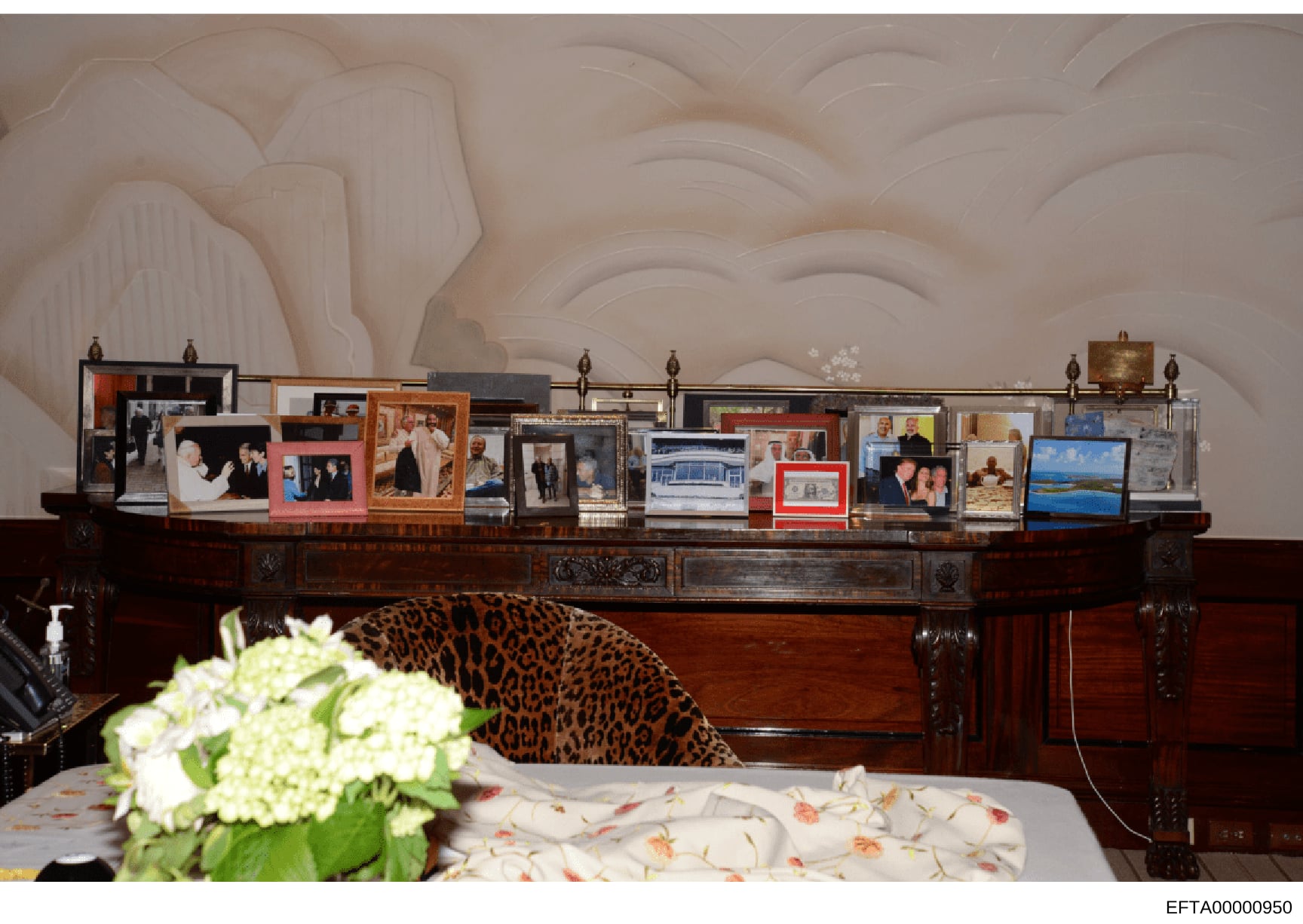 This photograph documents the interior of a bedroom, featuring a dark wood mantelpiece displaying approximately 20 framed photographs, decorative items, and what appears to be landscape artwork. The scene includes ornate ceiling details, a bed with f
