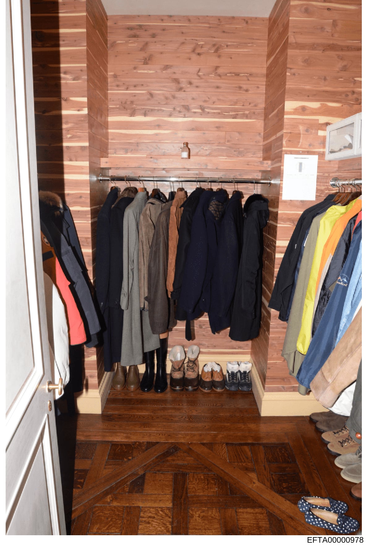 This is a photograph of an interior closet space featuring wooden paneled walls, a clothing rod with numerous hanging garments, and shoes arranged on the floor. The image shows various coats, jackets, and outerwear in dark colors along with some colo