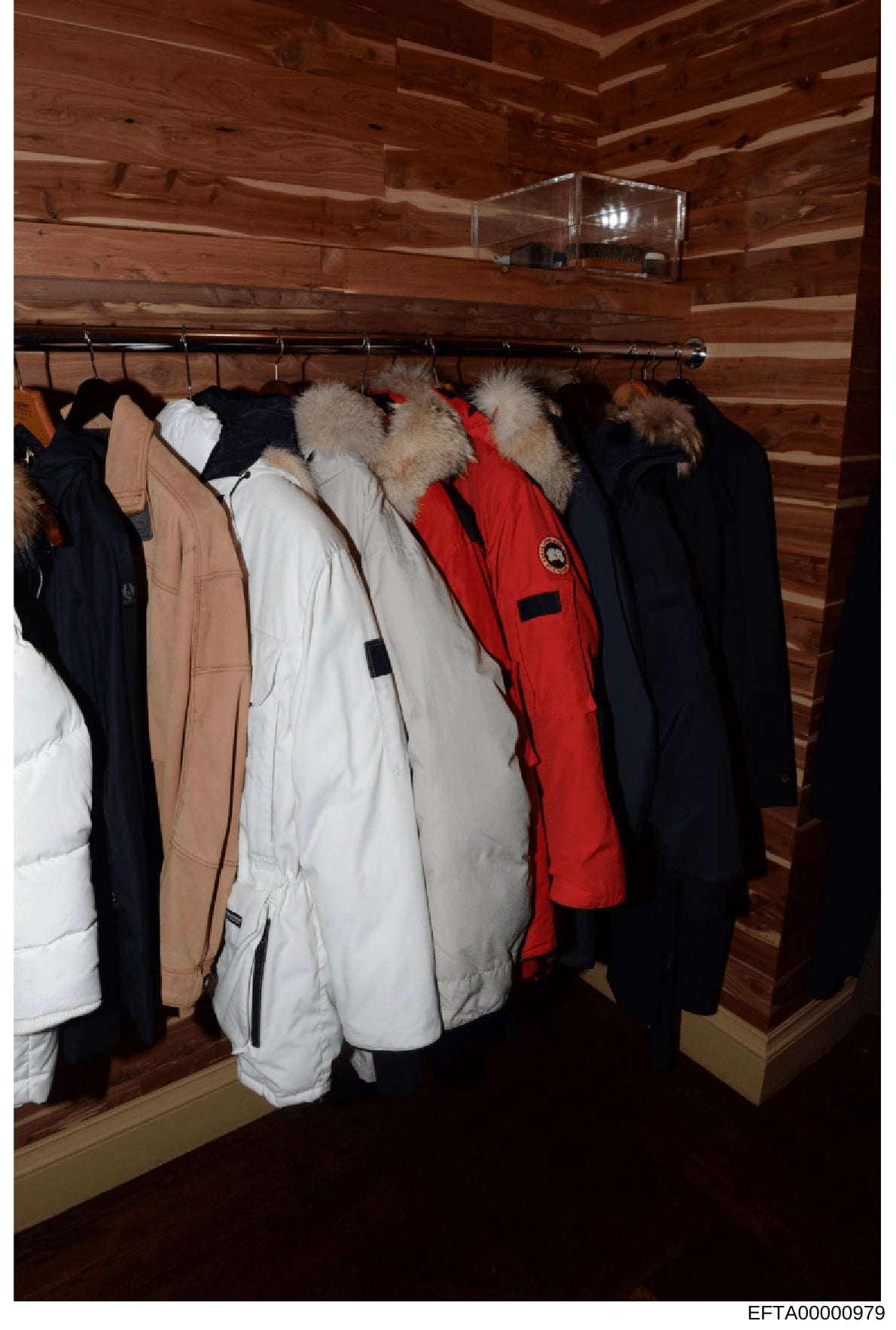 This photograph shows the interior of a wooden-paneled closet containing multiple winter coats hanging on a metal rod. The collection includes fur-trimmed parkas in various colors (navy, tan, white, red, and black), with several featuring coyote or s