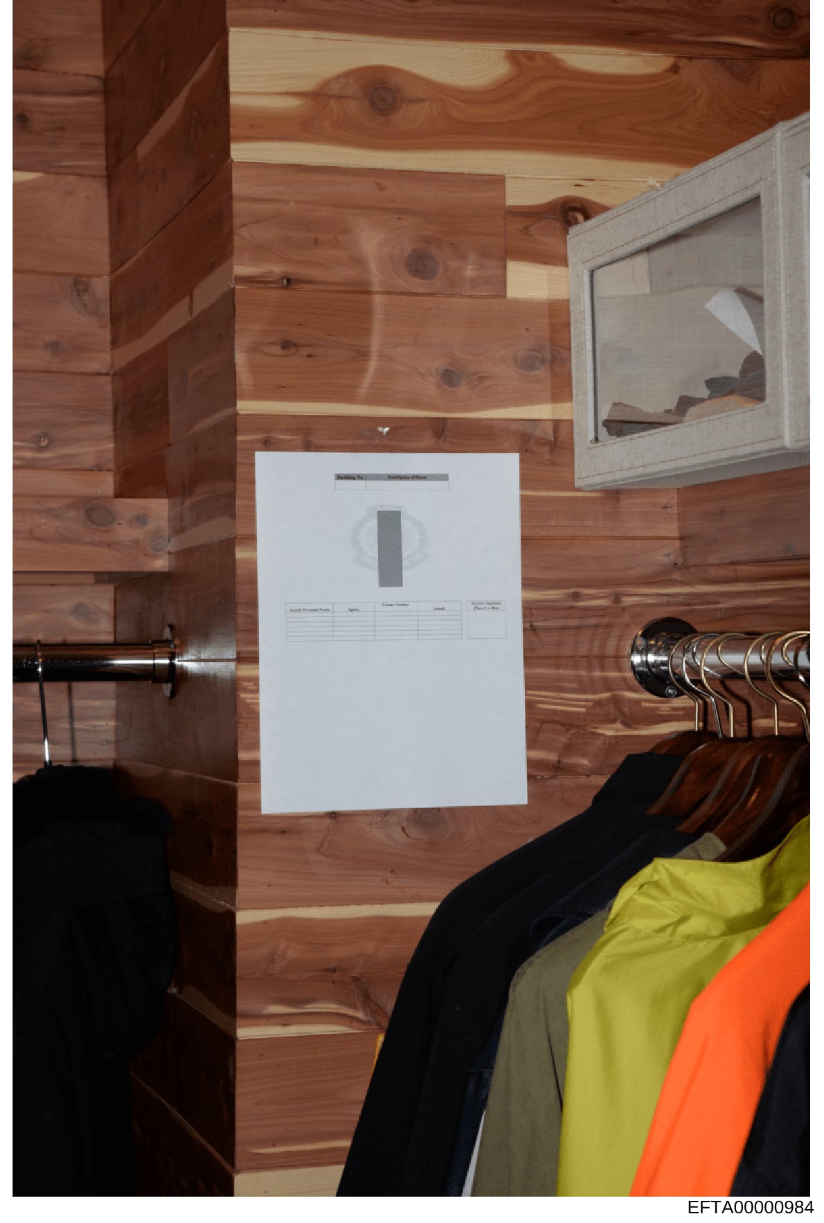 This is a forensic evidence photograph showing the interior of a wood-paneled room with a closet, clothing on hangers, and an evidence collection marker placed on the wall. The marker appears to be used to document and scale evidence during an invest