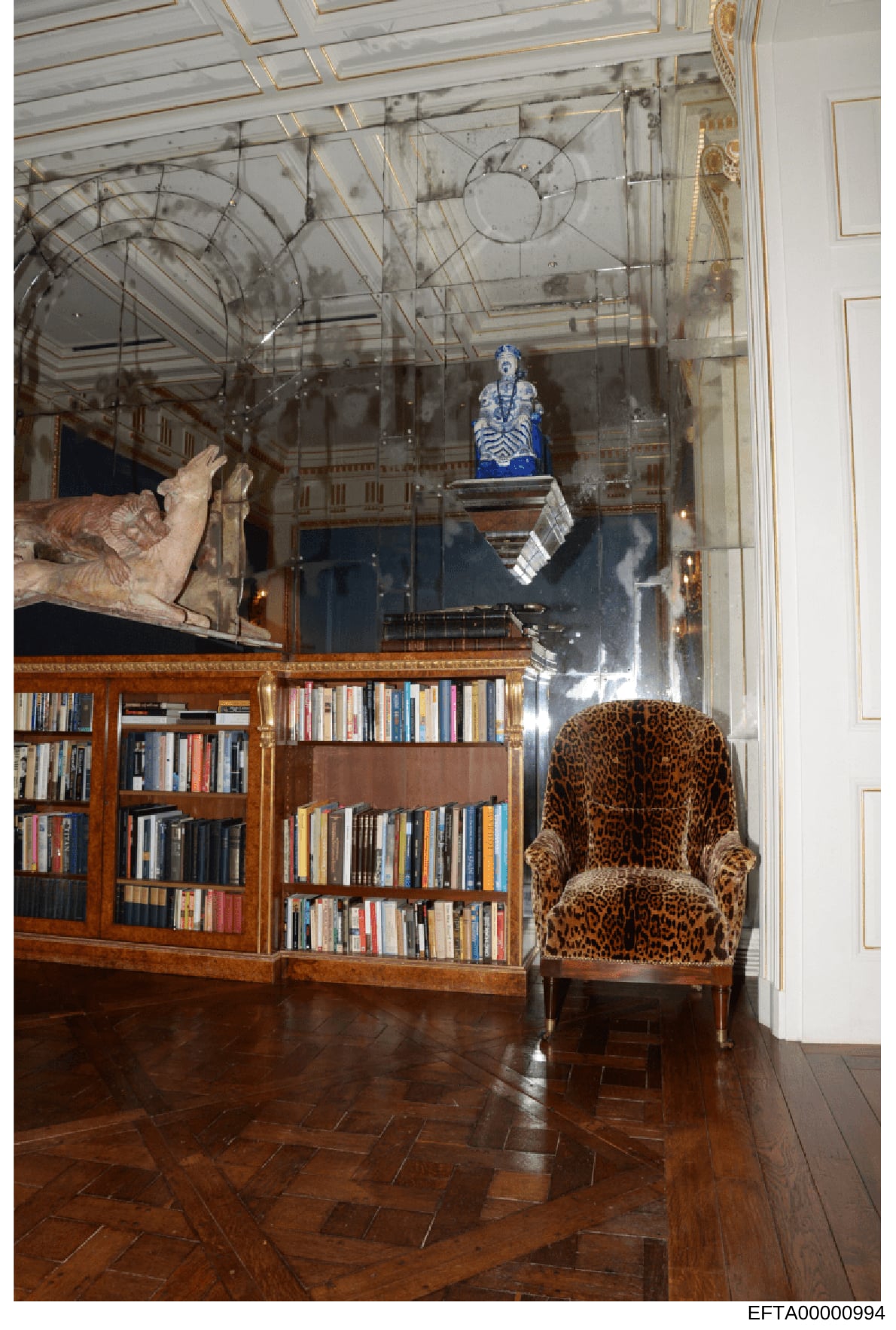 This photograph documents an elegantly appointed library or study room featuring high ceilings with ornate architectural detailing, built-in wooden bookshelves, fine art objects, and luxury furnishings. The room exhibits classical interior design wit