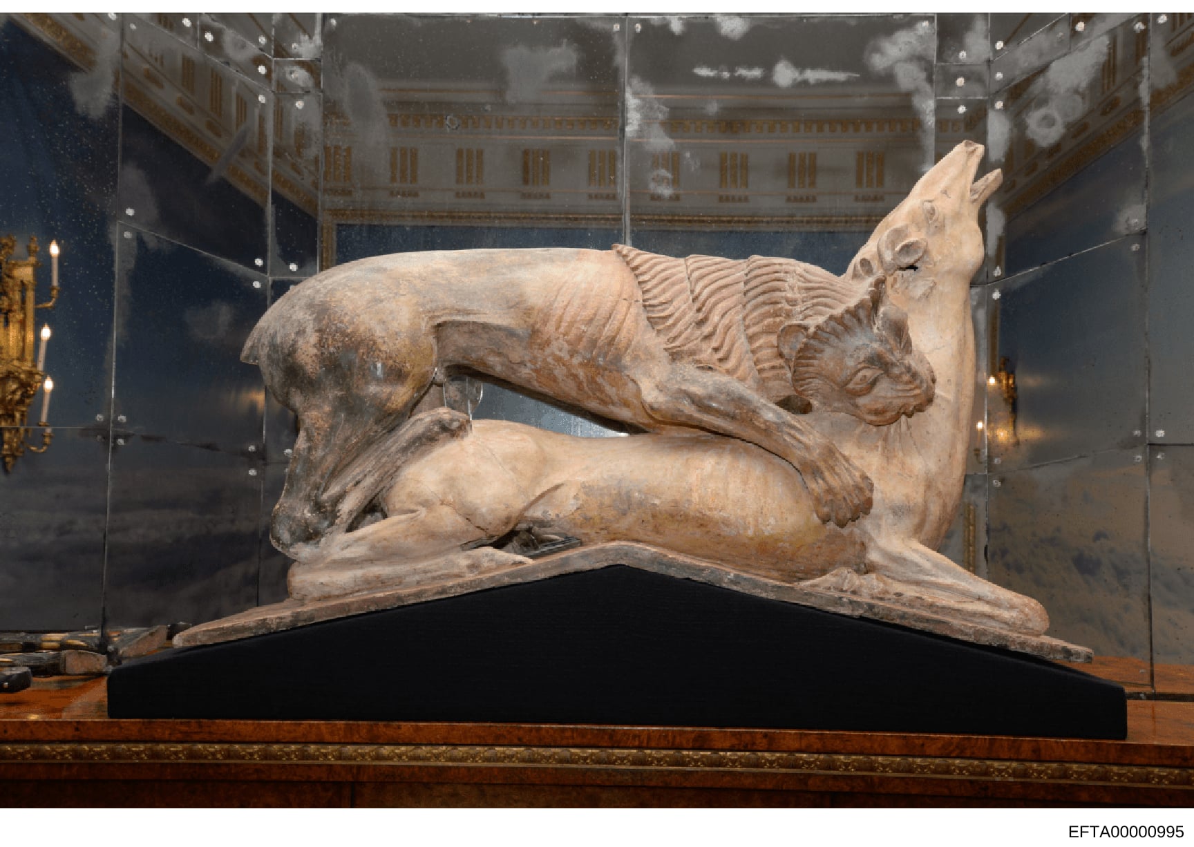 This photograph documents a classical or neoclassical stone sculpture depicting a reclining mythological creature (possibly a dragon or chimera) displayed in a museum-quality glass case. The artifact is mounted on a dark base within a climate-control