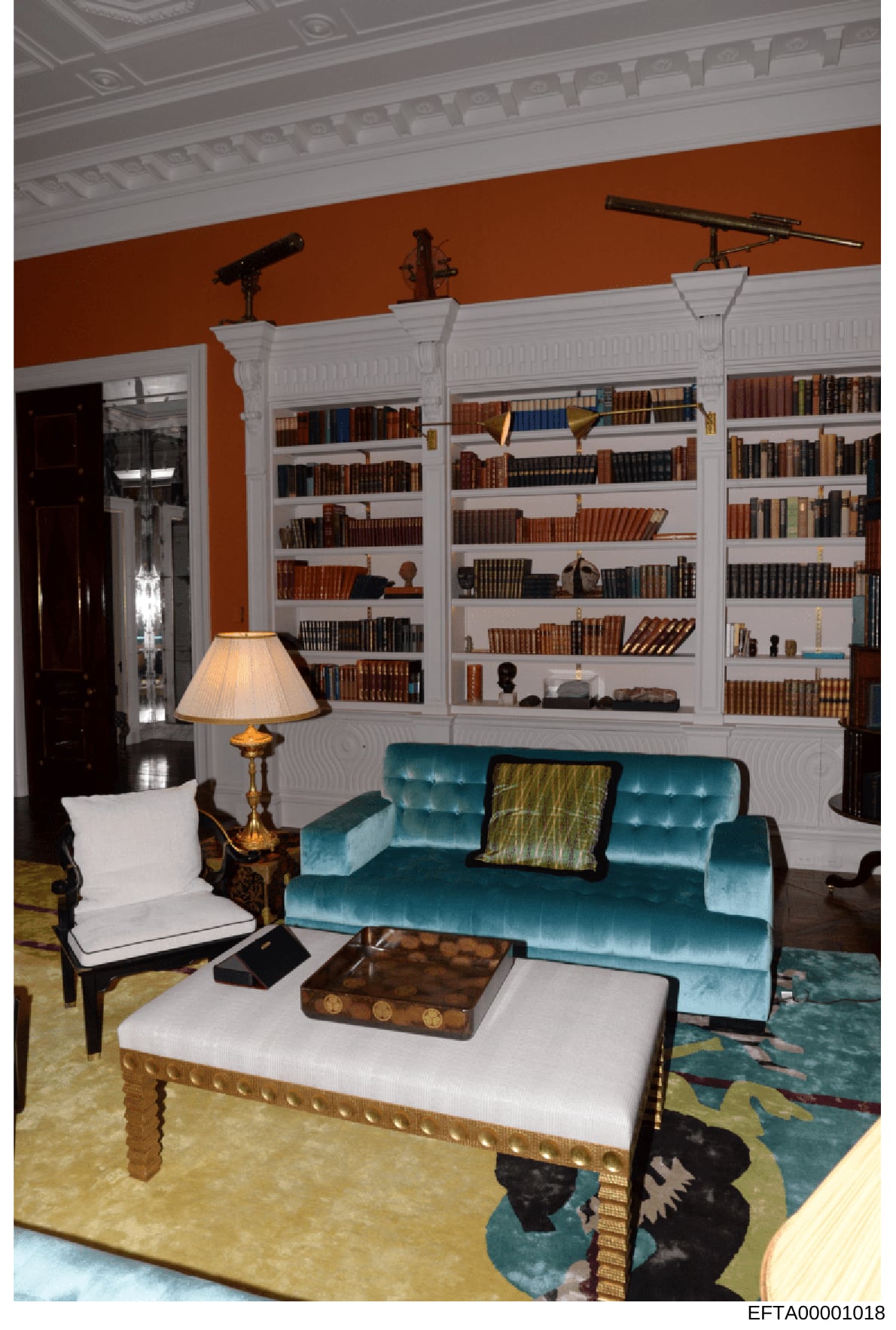 This photograph shows an elegantly appointed library or study room featuring floor-to-ceiling white built-in bookshelves filled with leather-bound books, a turquoise velvet sofa with decorative pillows, ornamental tables, and period furnishings. The 