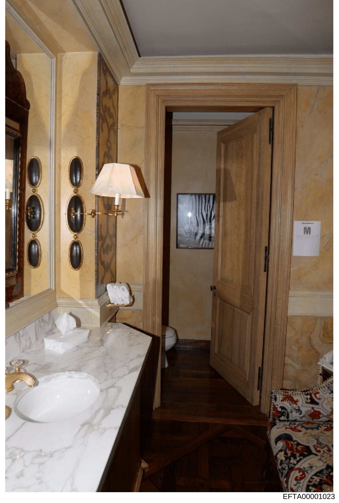 This is a photograph of an upscale bathroom interior, likely from one of Jeffrey Epstein's properties. The image shows high-end finishes including marble countertops, gold-plated fixtures, ornate wall sconces, and custom cabinetry. The bathroom is la