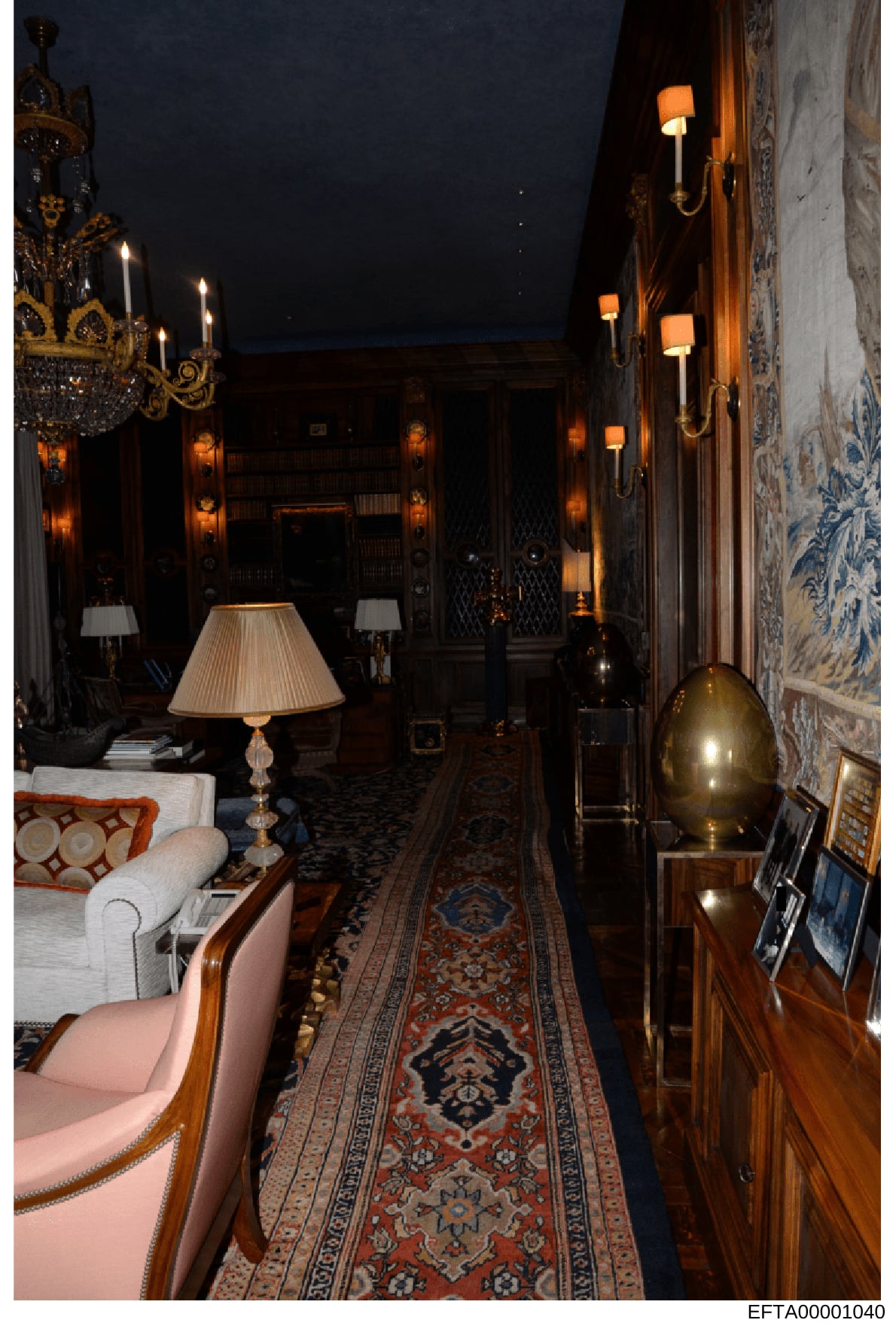 This is a photograph of an elegant interior hallway or gallery featuring high-end furnishings and décor. The space displays ornate chandeliers, wall sconces, decorative furniture including upholstered chairs, fine rugs, and carefully curated lighting