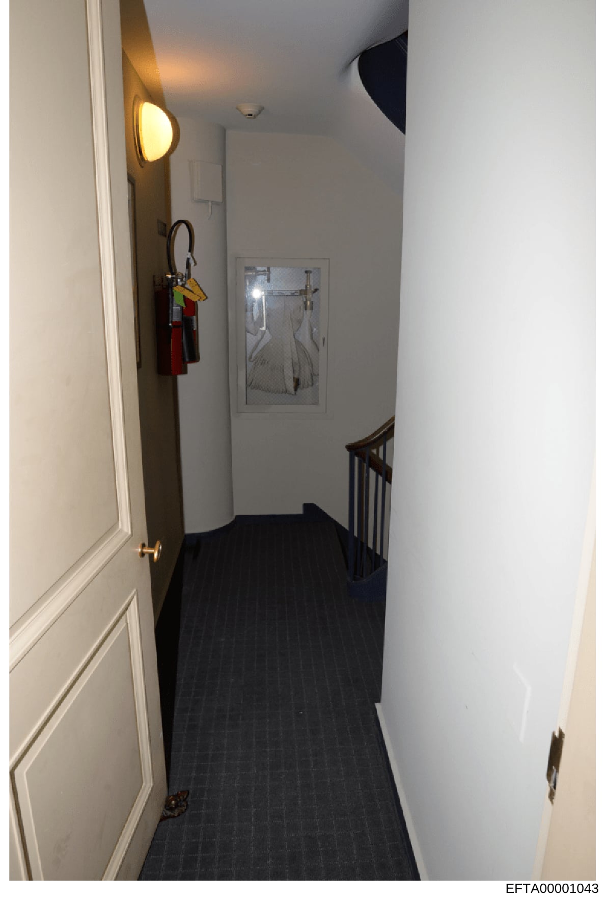 This is a photograph of an interior hallway within a residential property, showing a narrow corridor with white walls, dark flooring, decorative sconce lighting, framed artwork, and a staircase. The image appears to be documentation of the interior o