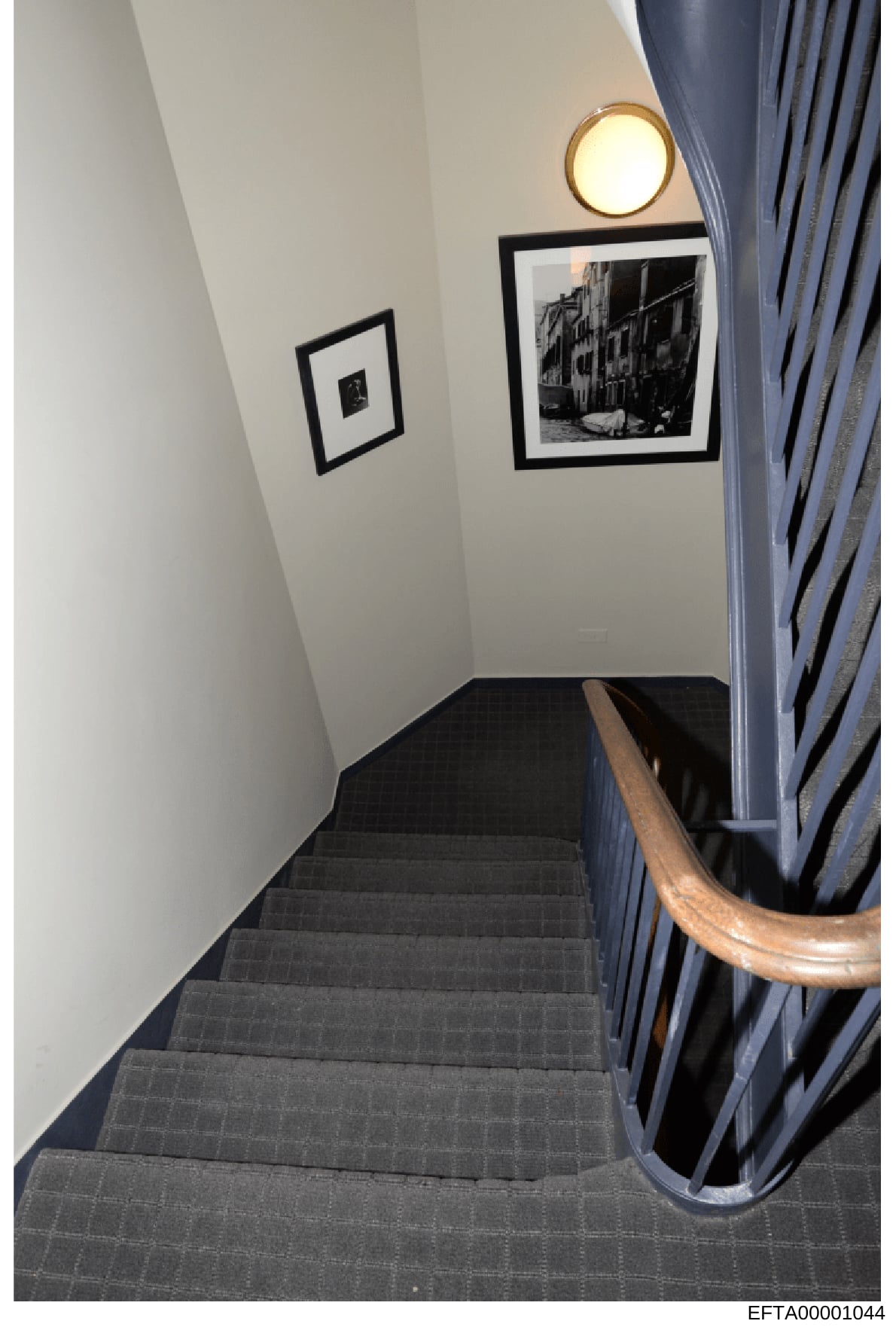 This is a photograph of an interior stairwell in what appears to be a residential or commercial property. The image shows a narrow staircase with dark gray carpeting, white walls, framed black-and-white artwork, a round wall sconce, and blue curtains