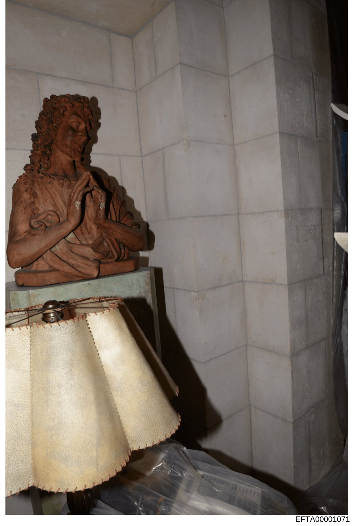 This is a photograph of an interior space showing a terracotta or brown-colored decorative bust sculpture positioned on a pedestal or furniture piece with a cream-colored fabric skirt. The background features white subway tile walls. The image appear