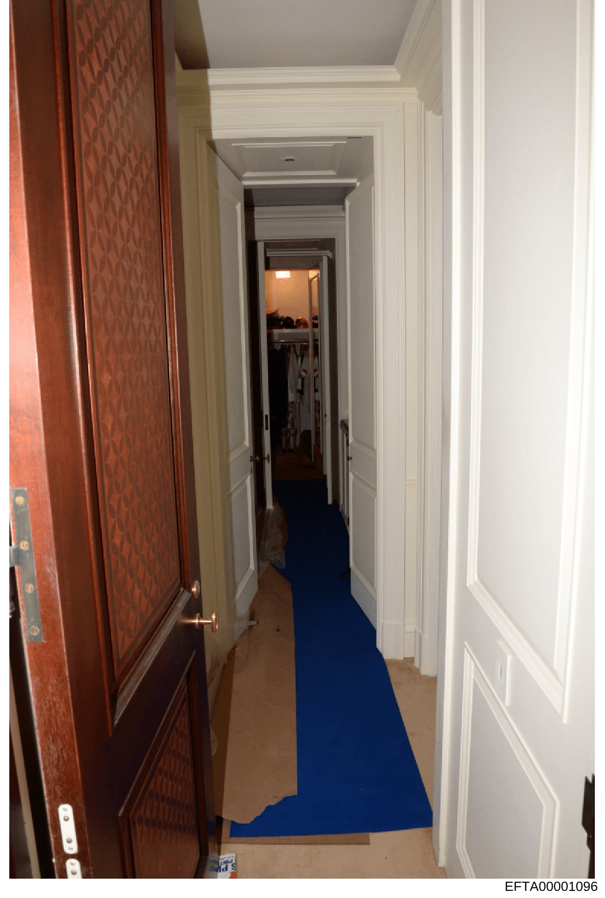 This is a photograph of a long, narrow hallway in what appears to be a residential or commercial property. The hallway features white paneled walls, recessed lighting, cream and dark wood trim, and a distinctive blue runner carpet down the center of