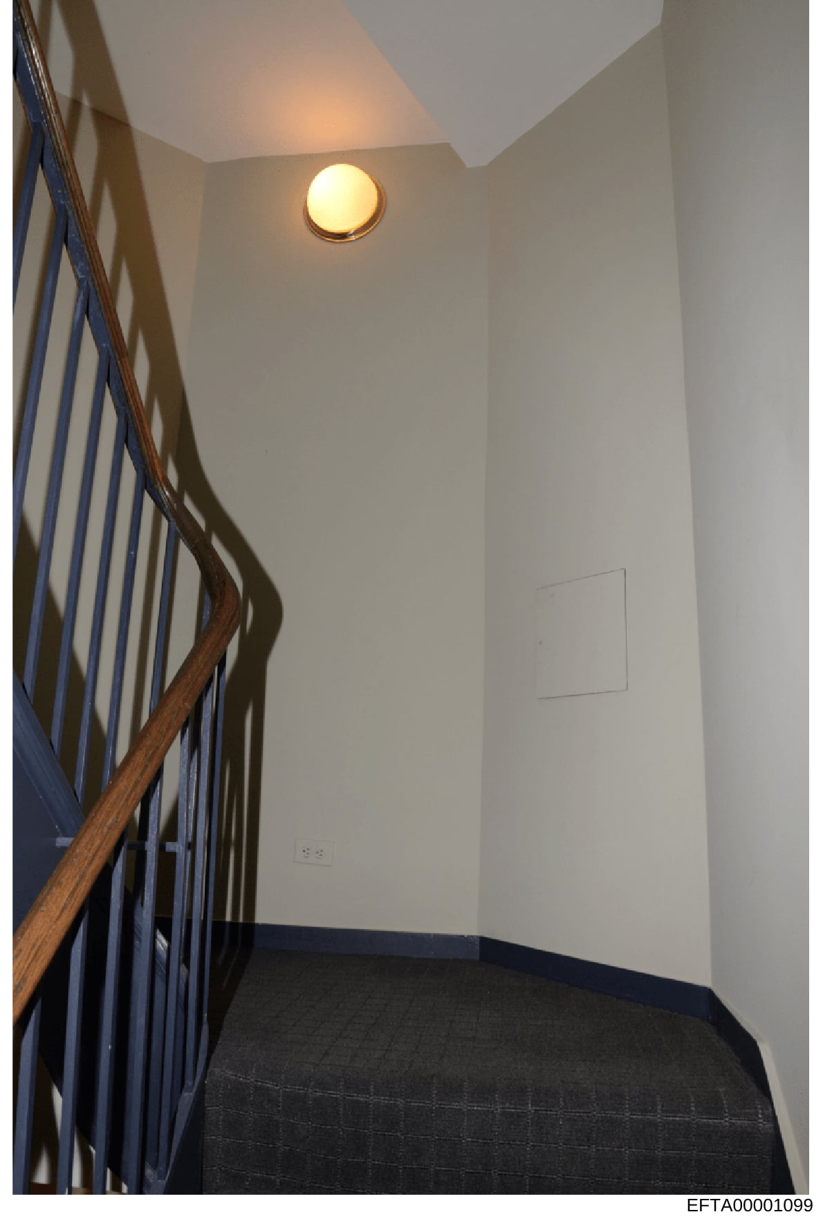 This is a photograph of an interior stairwell or landing area, showing white painted walls, a wooden stair railing with metal spindles, recessed ceiling lighting, and dark flooring. The image appears to be documentation of a residential or commercial
