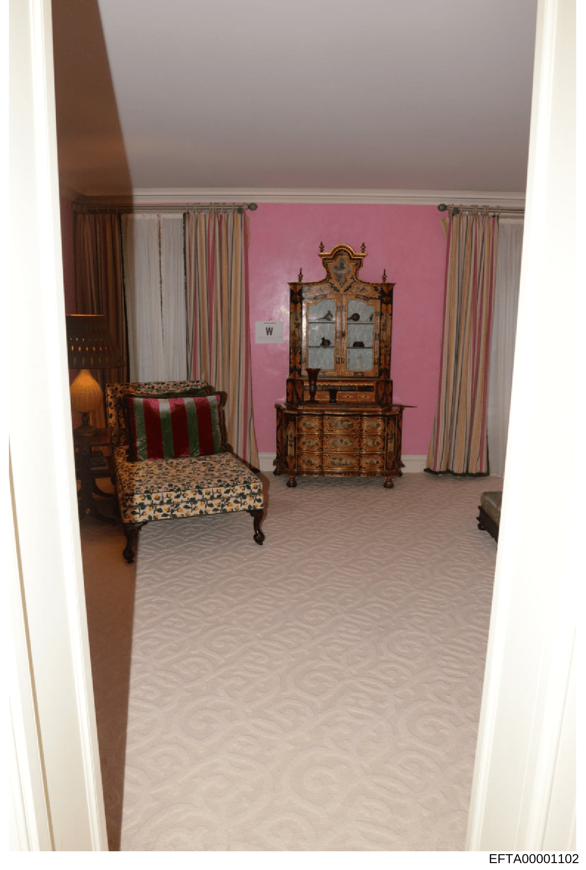 This is a photograph of an interior bedroom space featuring pink walls, antique furnishings, and decorative elements. The room contains a patterned upholstered chair, an ornate wooden cabinet with glass doors, striped curtains, and period furniture c