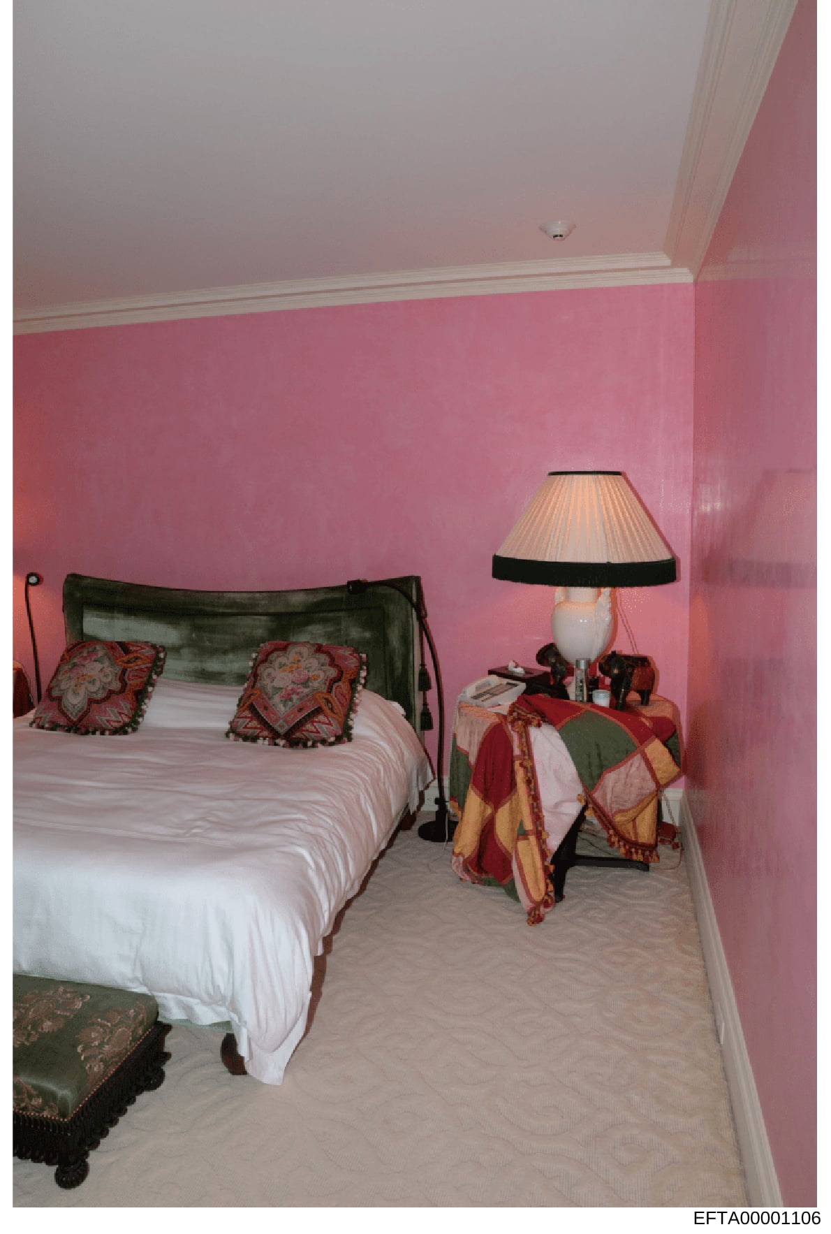 This is a photograph of a residential bedroom featuring pink-painted walls, a metal-framed bed with decorative red and gold pillows, a bedside table with lamp, and various furnishings. The image appears to be documentation of a property interior, lik