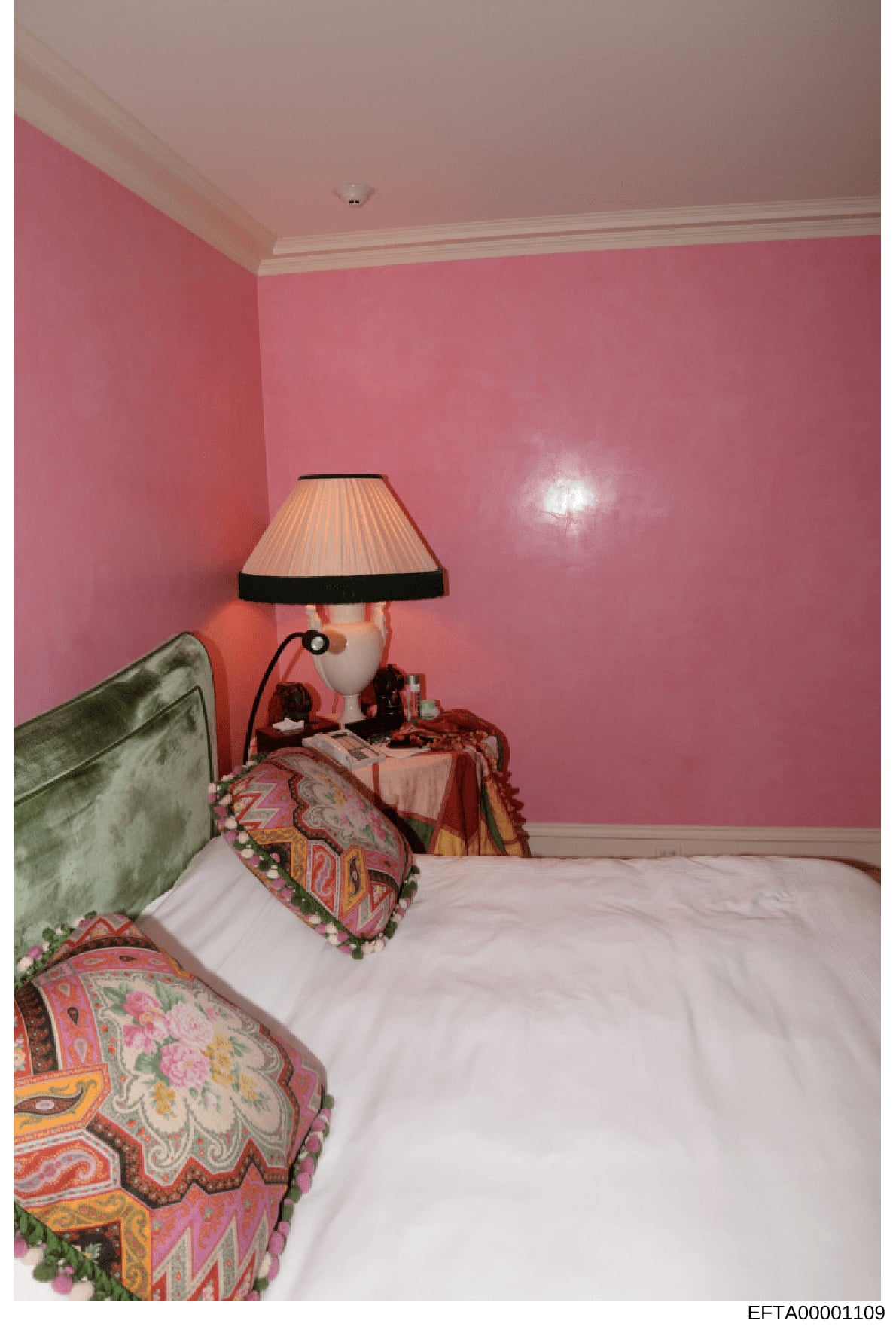 This is an evidence photograph showing the interior of a bedroom, featuring pink walls, a bed with colorful patterned pillows, a nightstand with a table lamp, and various decorative items. The photo appears to be part of property documentation from t