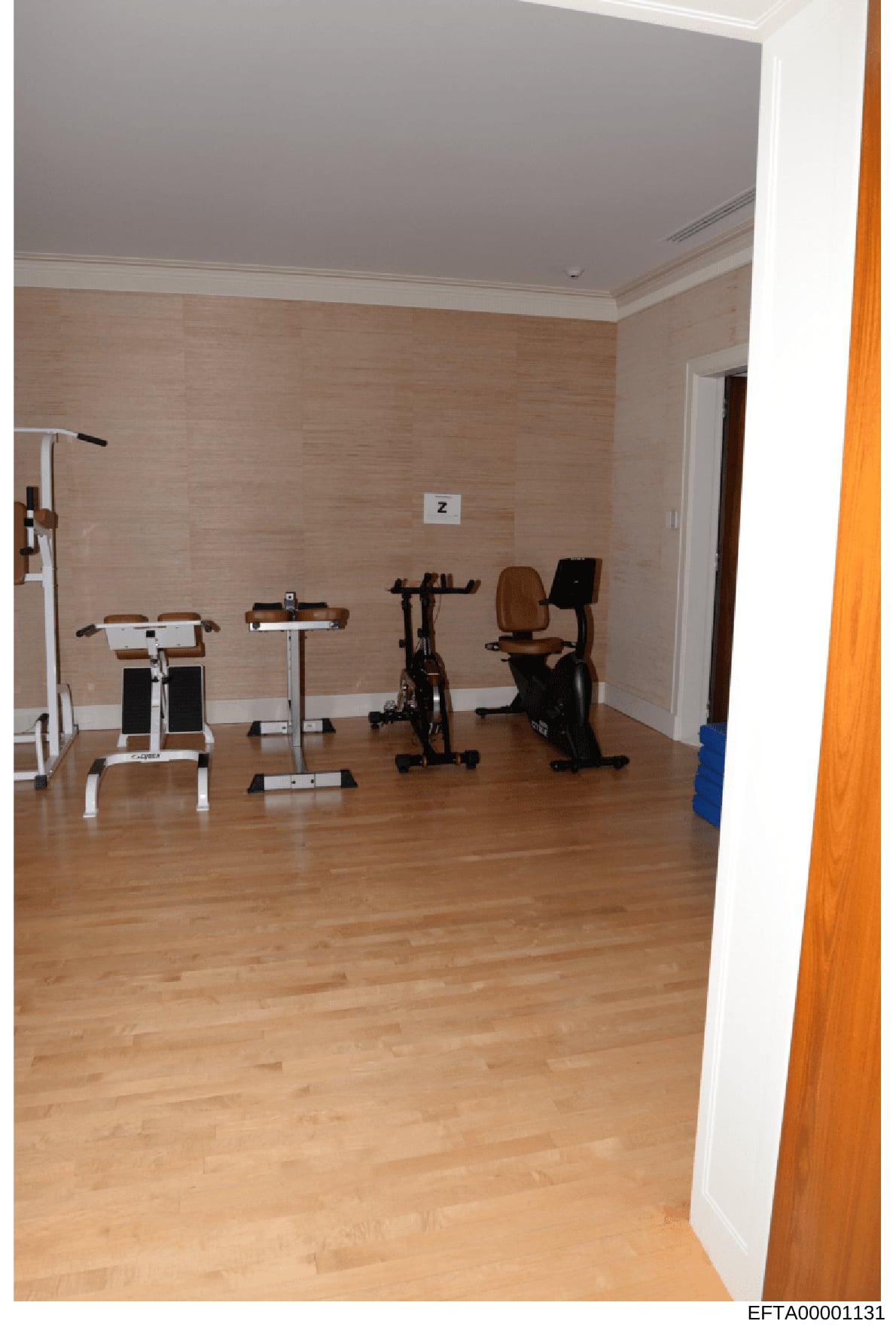 This photograph documents a residential gym space containing various exercise equipment including treadmills, stationary bikes, and other fitness machines. The room features light wood flooring, tan/beige textured walls, and white crown molding. This
