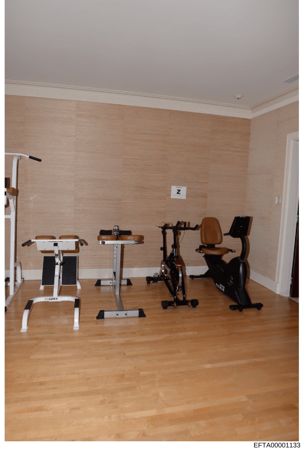 This is a photograph of an indoor home gym or fitness room containing multiple pieces of exercise equipment arranged along a wall. The room features wooden flooring, beige textured wallpaper, and contains at least four different exercise machines inc