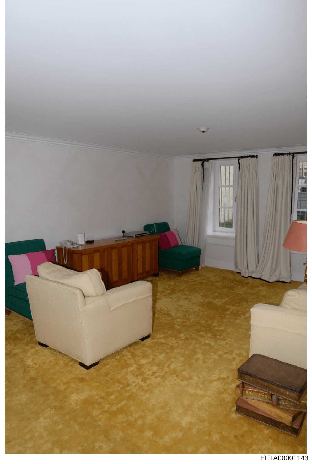Photograph showing the interior of a living room with cream-colored walls, beige carpet, and mixed furnishings including cream upholstered chairs, green seating, a wooden desk, and cream curtains. The room appears to be part of a larger residential p