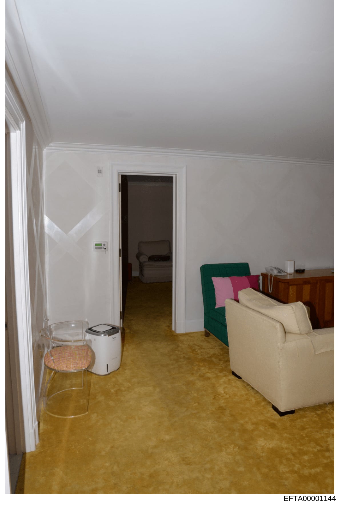This is a photograph of an interior room, likely a bedroom or sitting area, showing white walls, cork or tile flooring, a cream-colored sofa with green and pink throw pillows, a wooden dresser, and a doorway leading to an adjacent room. The image app