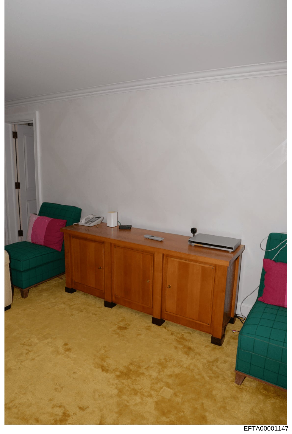 This is an evidence photograph showing the interior of a room, likely from one of Epstein's properties. The image depicts a furnished space with a wooden desk/cabinet, green upholstered chairs with pink/red cushions, white walls with crown molding, a