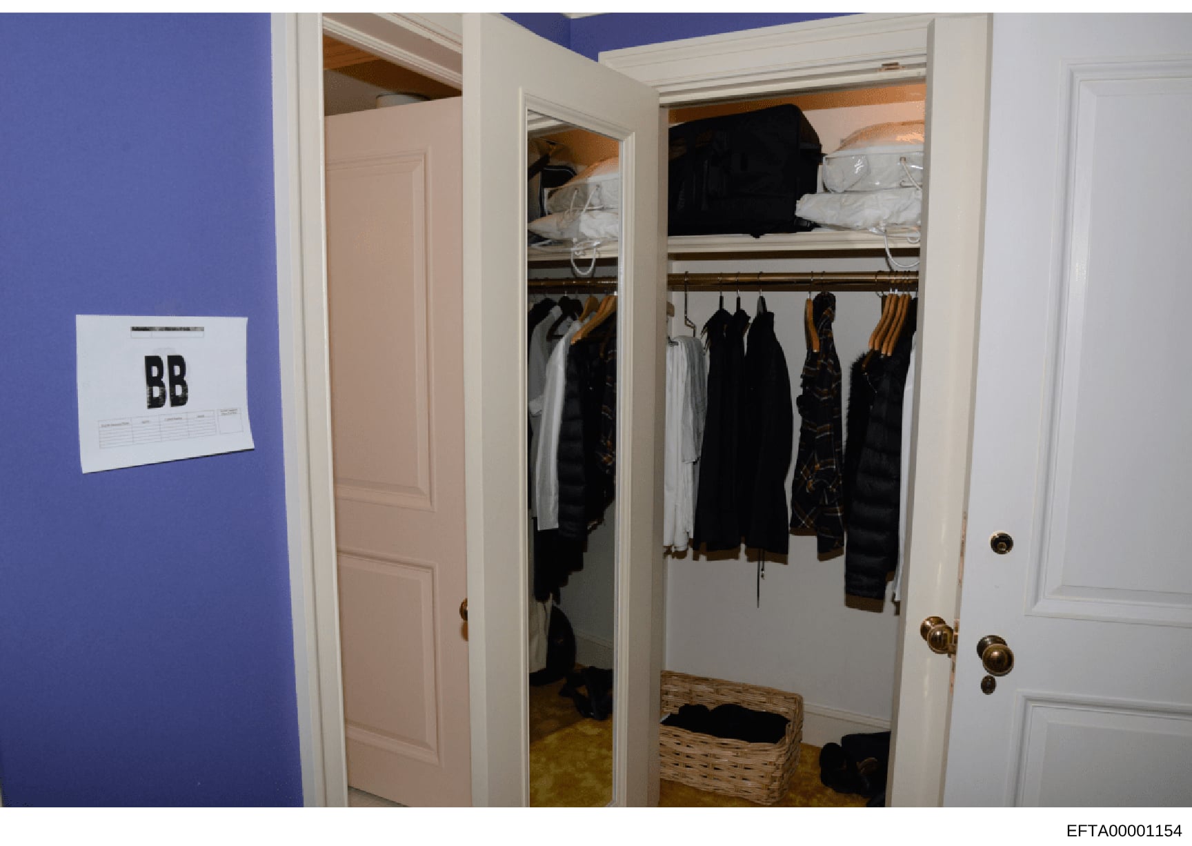 This is a photograph of a residential walk-in closet labeled 'BB', likely evidence documentation from the Epstein investigation. The image shows clothing storage including multiple black garments hanging on rods, shelving with folded items and beddin