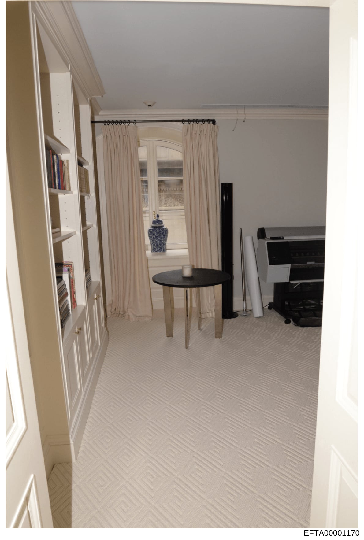 This is a photograph of an interior room, likely a bedroom or sitting area from one of Jeffrey Epstein's properties. The image shows a modern, elegantly furnished space with cream-colored walls, built-in shelving, cream curtains, a round black side t