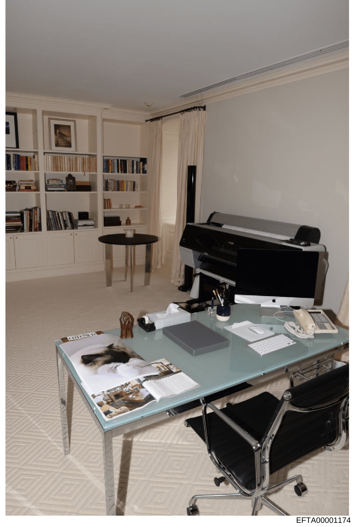 This is a photograph of an upscale residential office or study space, showing a modern work area with a glass desk, computer setup, and extensive built-in white bookshelves. The room features a black grand piano, round side table, and elegant furnish
