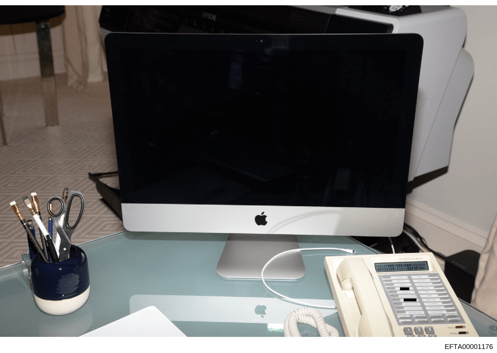 This photograph shows a contemporary office workspace featuring an Apple iMac desktop computer, wireless mouse, office telephone system, desk organizer with writing implements, and other standard office equipment on a light blue desk surface. The ima