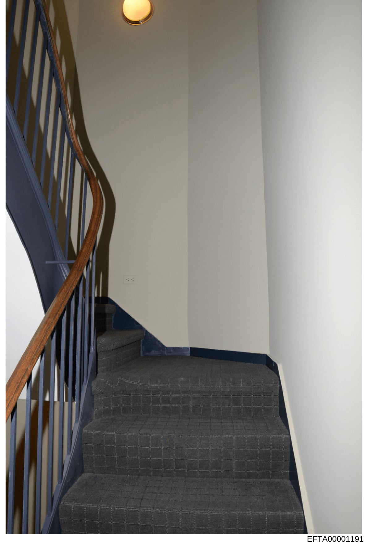 This photograph shows an interior staircase from one of the properties associated with the Epstein investigation. The image captures architectural details including a wooden handrail, metal spindles, dark-colored carpeting on the steps, white walls,
