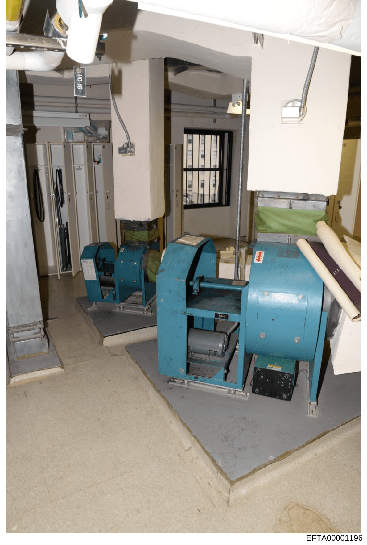 This photograph shows a basement or utility room containing two large blue industrial machines, electrical panels, storage racks, and various equipment. The space appears to be part of a residential or commercial property's mechanical systems area. T